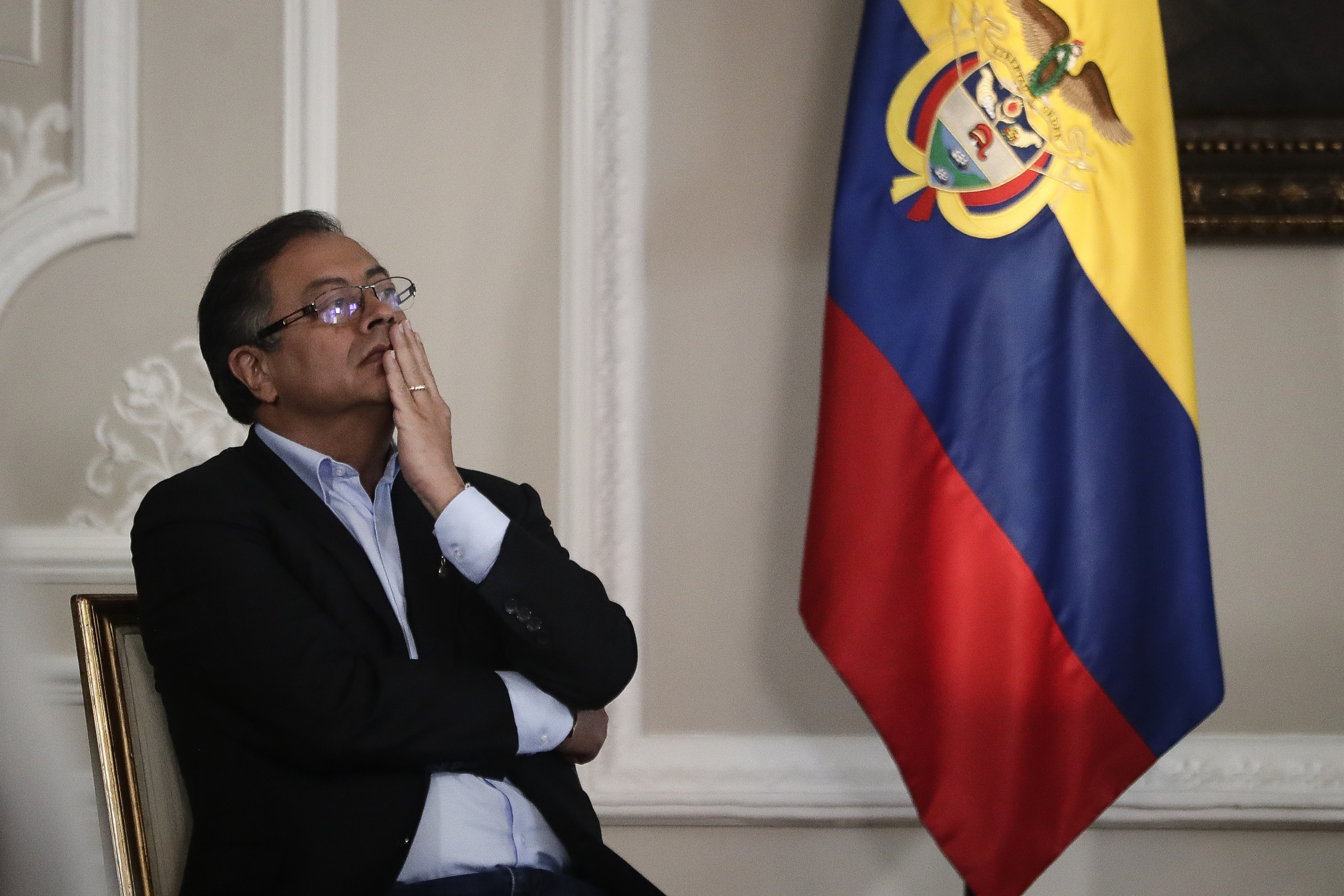 A man — Gustavo Petro — sits in a chair with arms folded and a hand on his face, as if lost in thought. A Colombian flag is next to him.