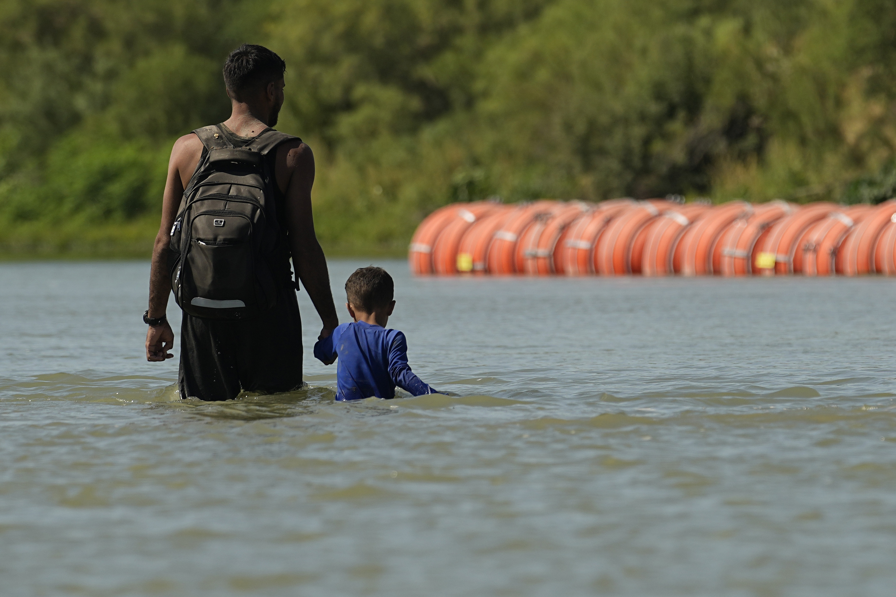 A migrant standing in the river stares at a floating border barrier