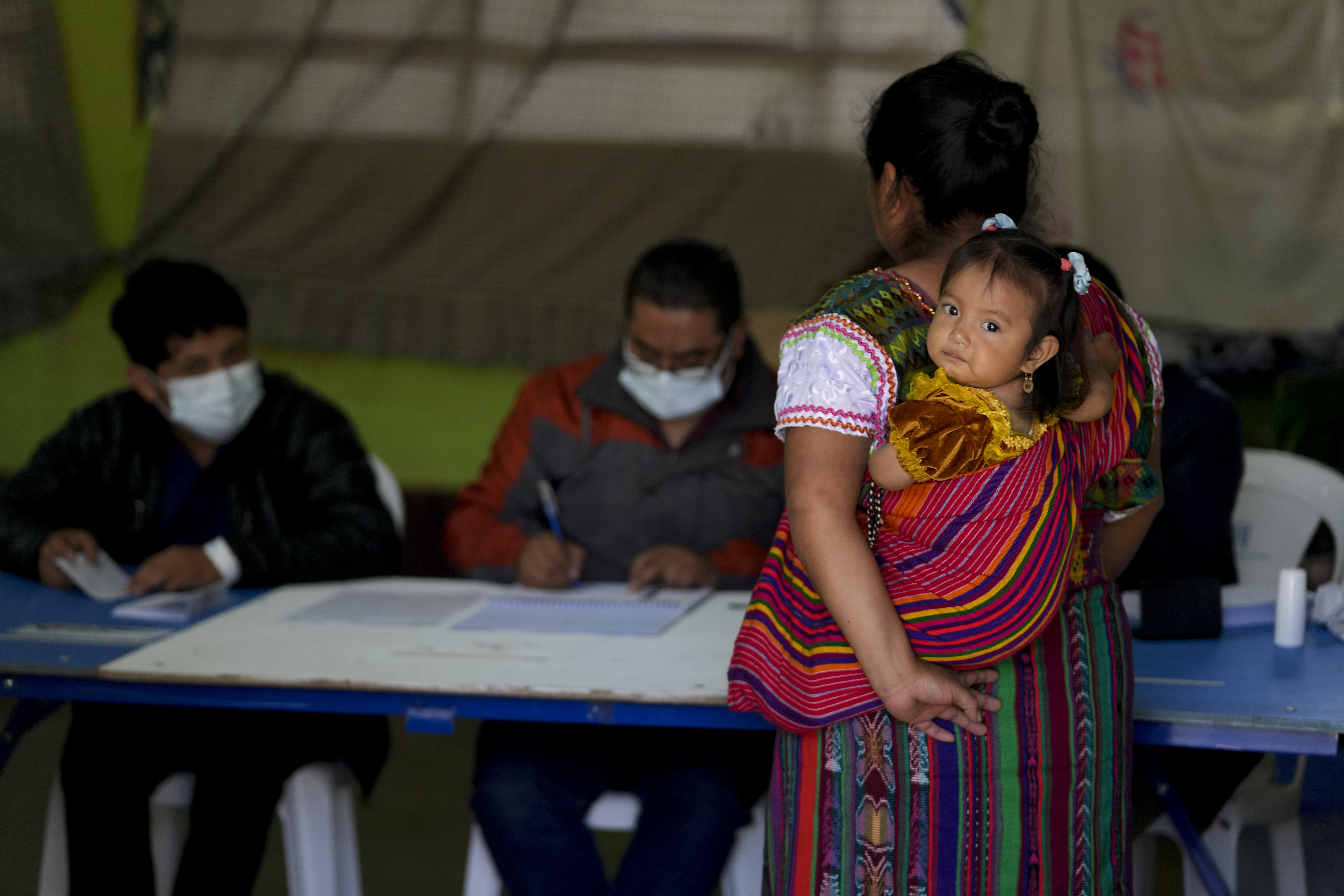 A woman arrives at a polling station. She is carrying her infant daughter.in a sling on her side.