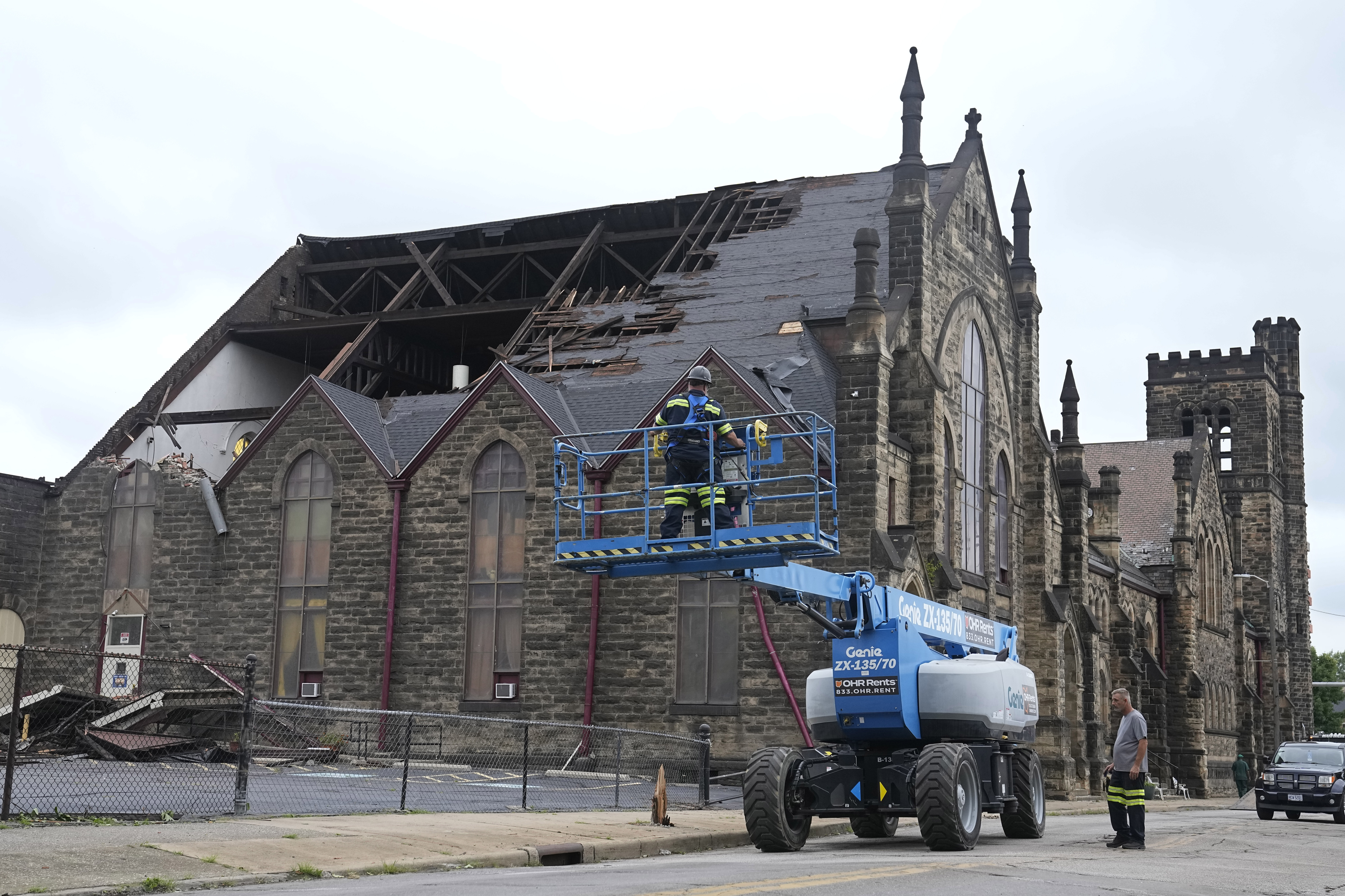 A Gothic-style church is seen with massive tears in its roof. A blue crane with workers in its basket is seen parked in the front.
