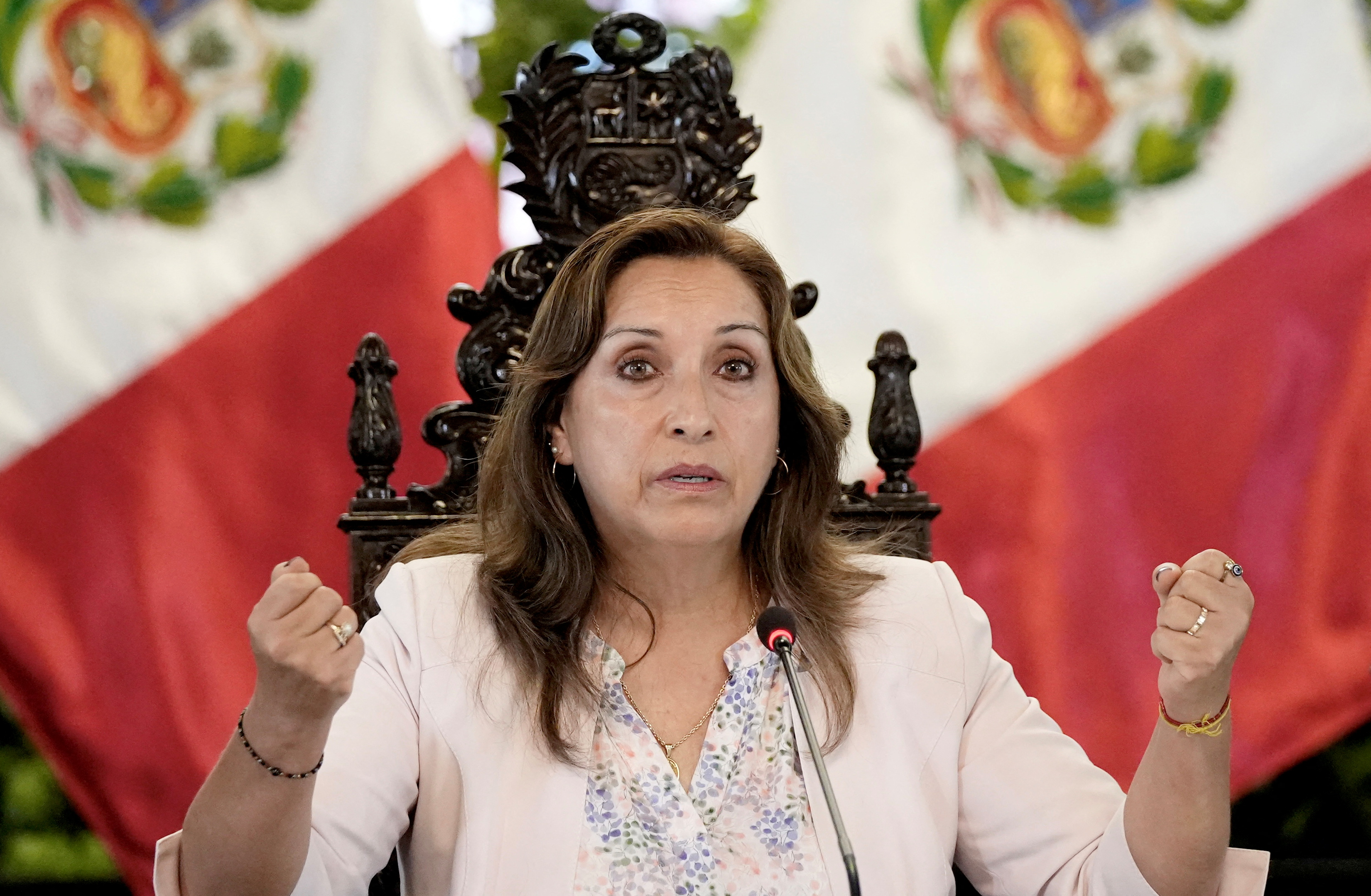 President Dina Boluarte, seated in a large wooden chair, holds up two firsts as she gestures and speaks into a microphone. Peruvian flags sit behind her.