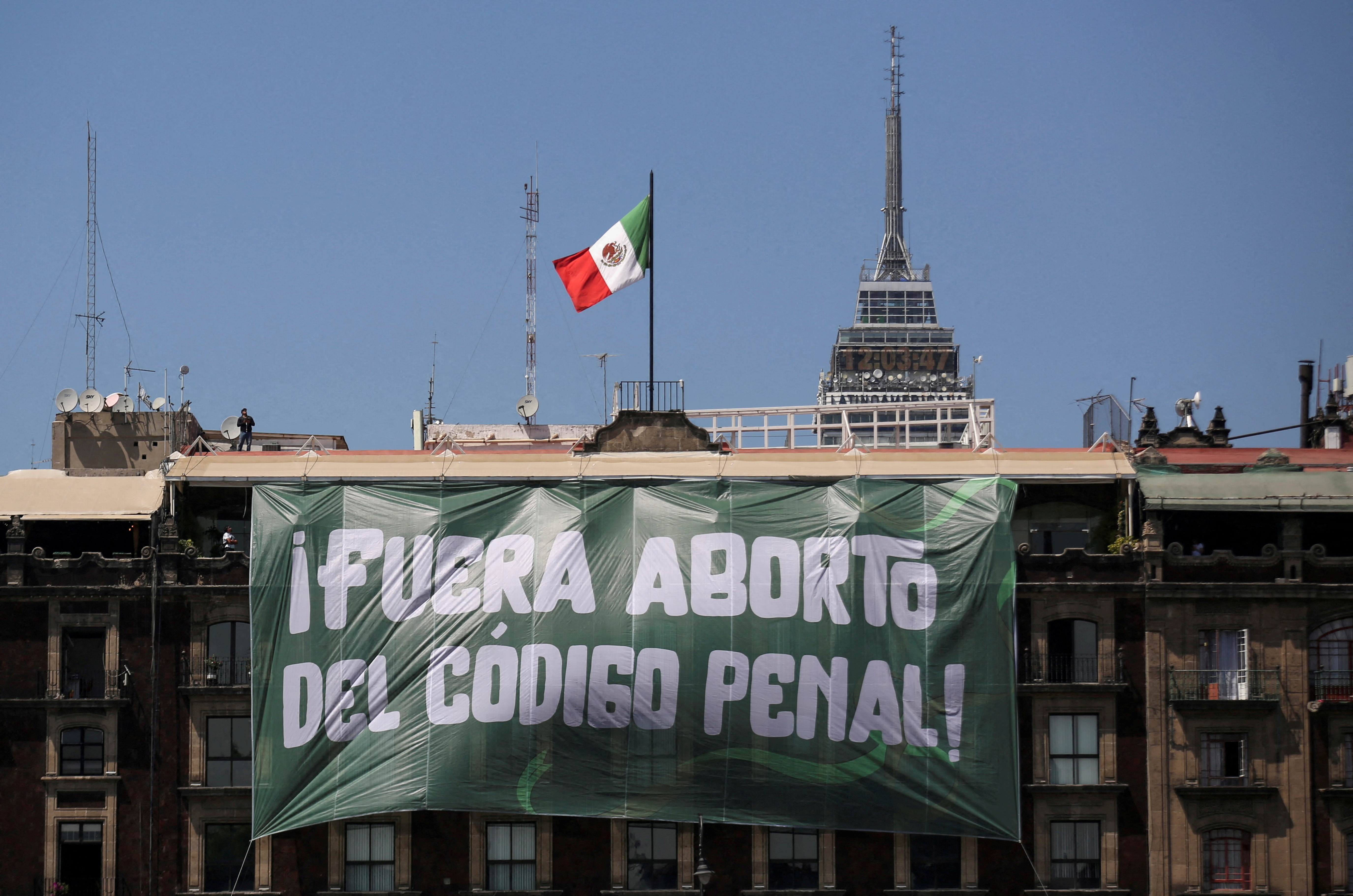 A green banner with white text hangs from a building topped by a Mexican flag.