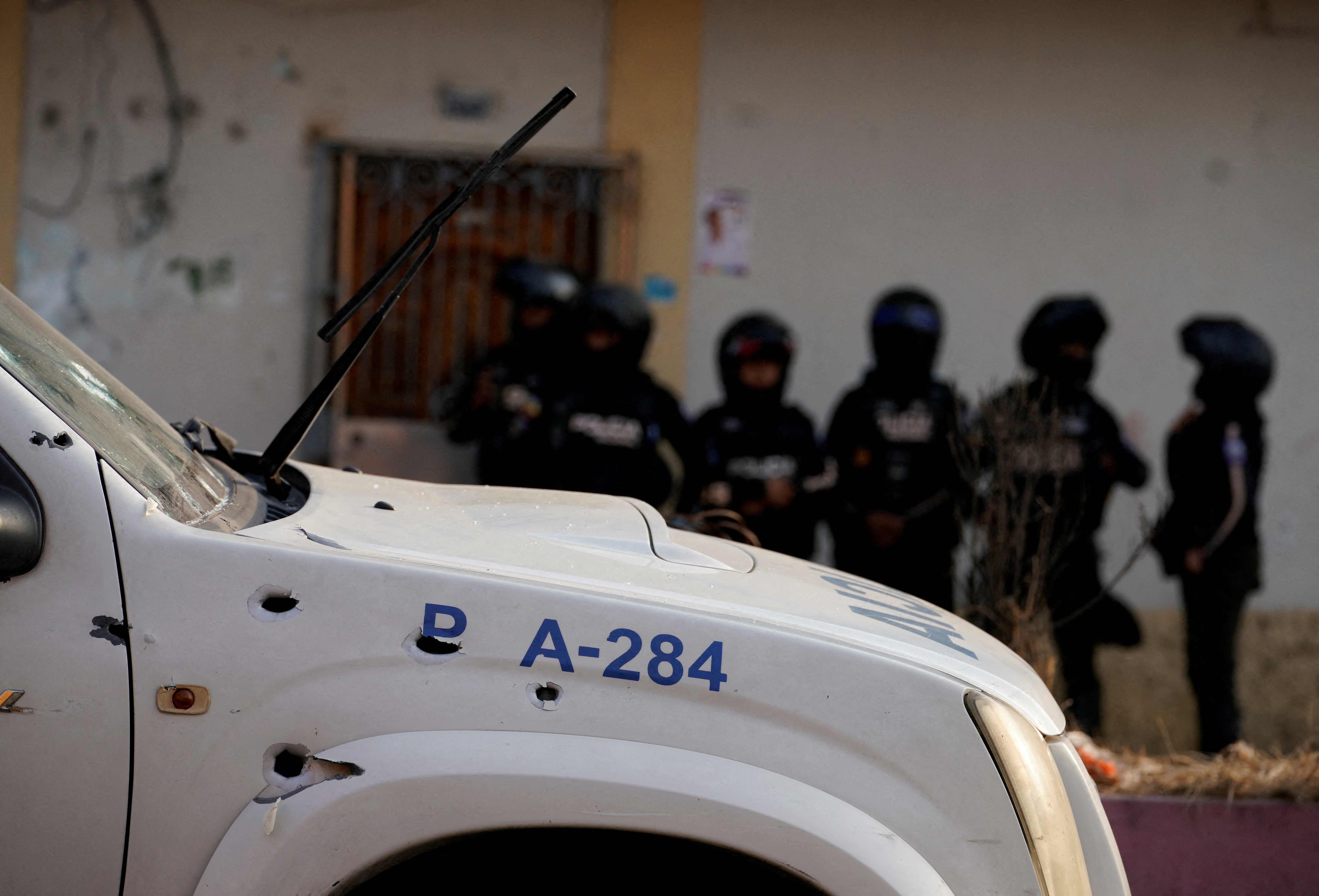 A white police car is parked on the road, riddled with bullet holes. Behind it, a line of officers dressed in black police tactical gear stand next to a building with an open door.