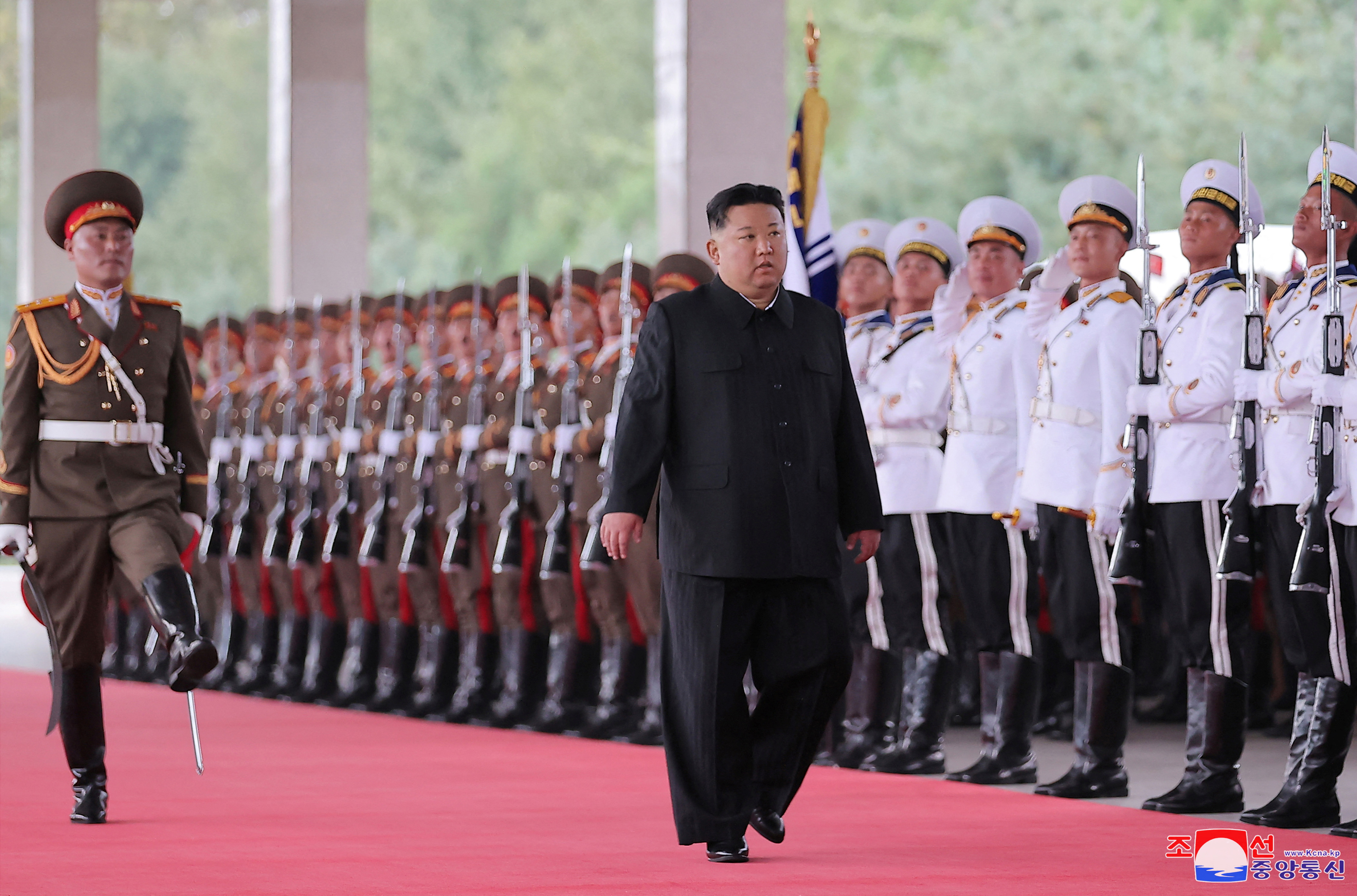 Kim Jong Un walking along a red carpet in front of a line of soldiers standing to attention. He is accompanied by another soldier who is goose stepping beside him