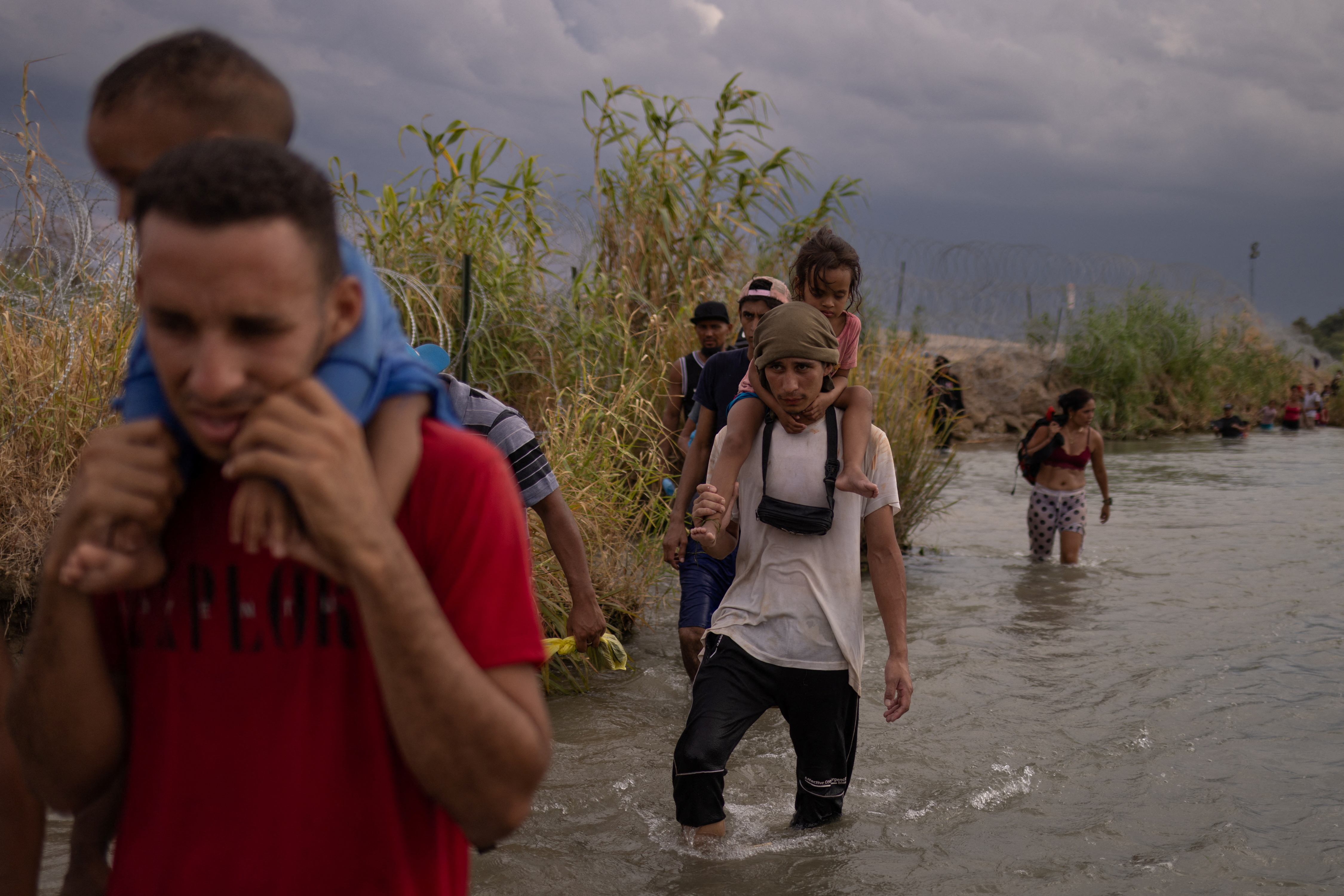 A man wades through muddy waters with a three-year-old on his shoulders, as others follow behind him, likewise carrying children or bags.