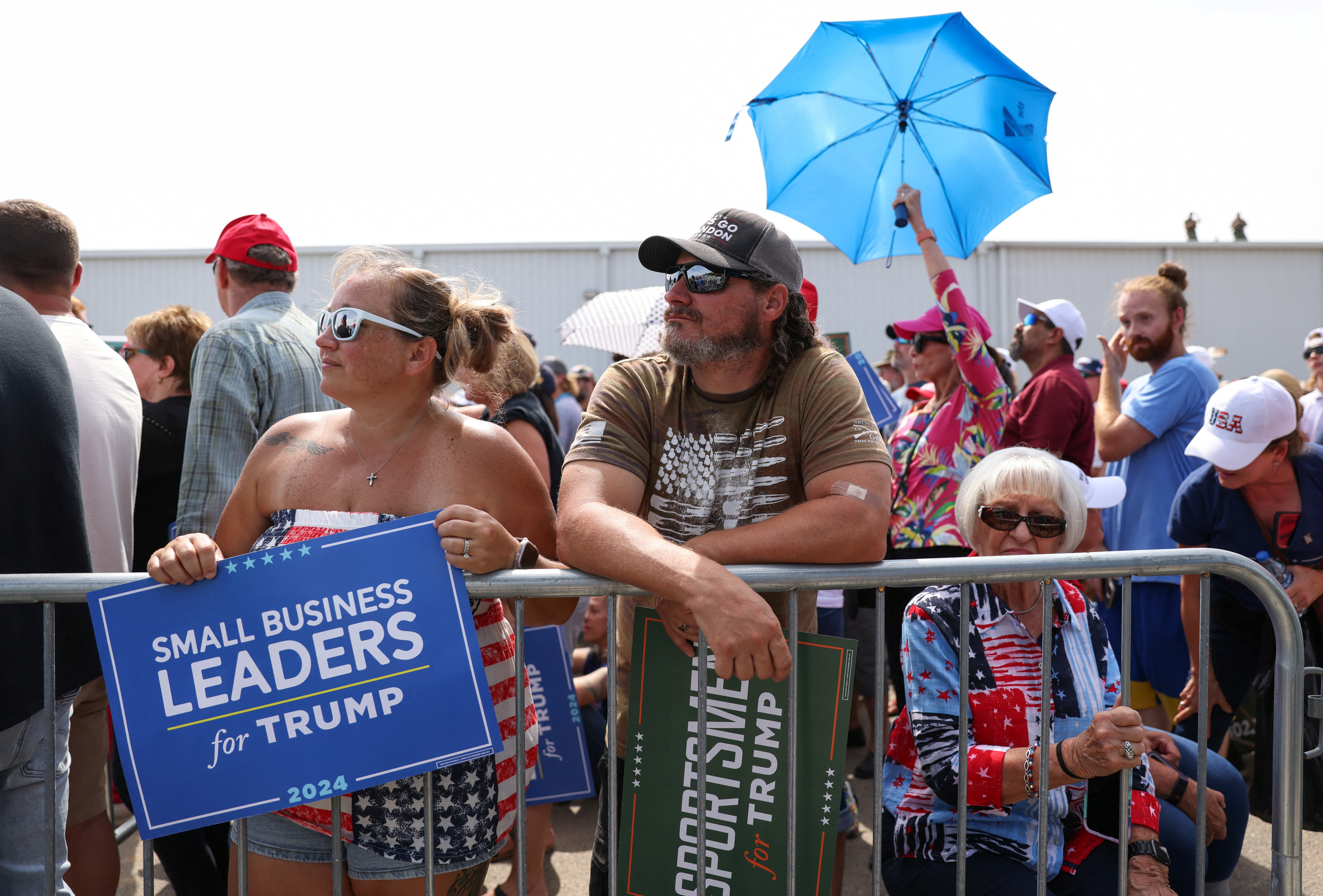 Supporters wait for former U.S. President and Republican presidential candidate Donald Trump during a 2024 presidential election campaign event at Sportsman Boats in Summerville, South Carolina, U.S. September 25, 2023. One person holds an umbrella. Another holds a sign that reads, "Small business leaders for Trump." Others lean against a railing to hold back the crowds.