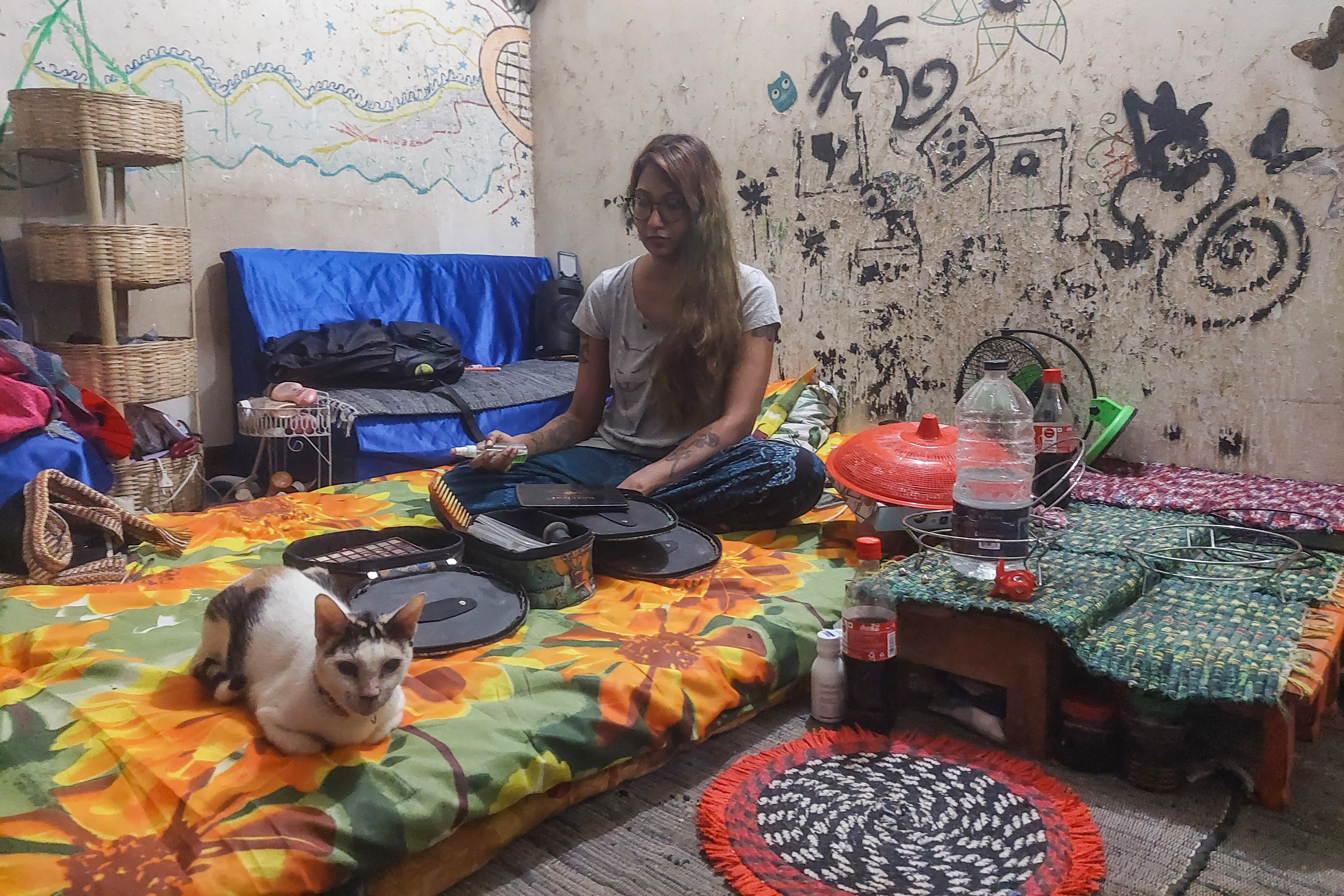 A photo of a woman sitting on a bed with a cat sitting in front of her while she looks through a bag.