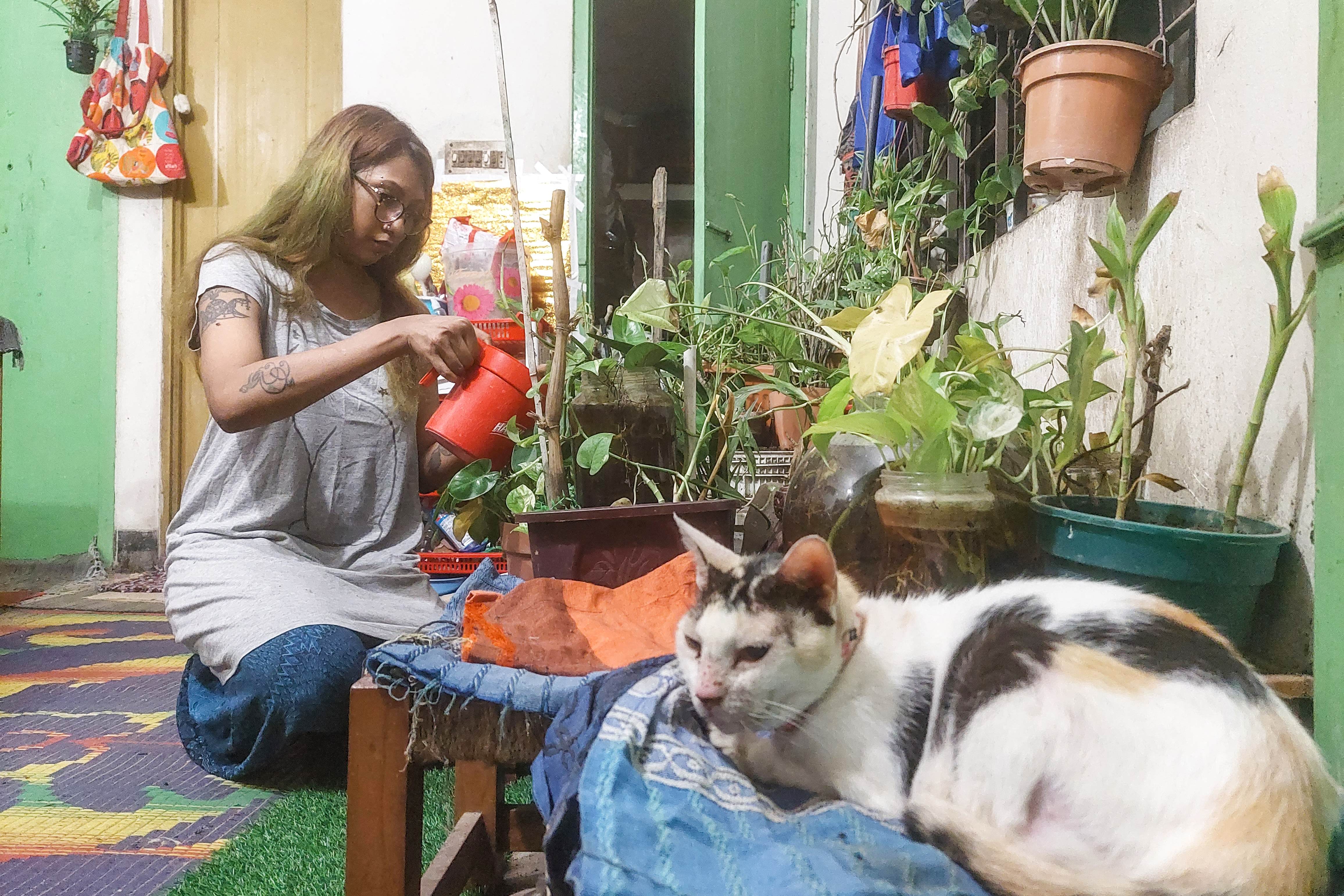 A photo of a woman watering some plants in pots on a table with a cat sitting on a table next to it.