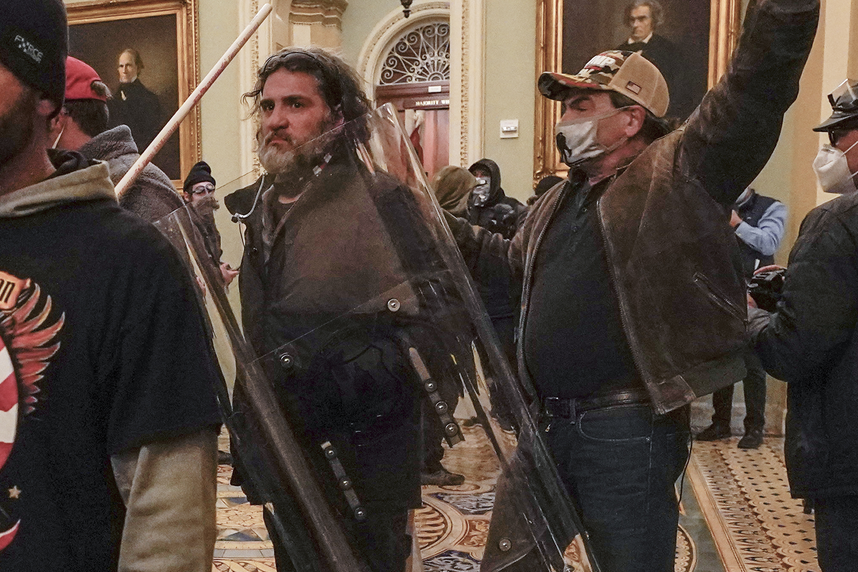 A man with a bushy beard carries a clear riot shield through the halls of the Capitol, as he and other rioters storm the building.