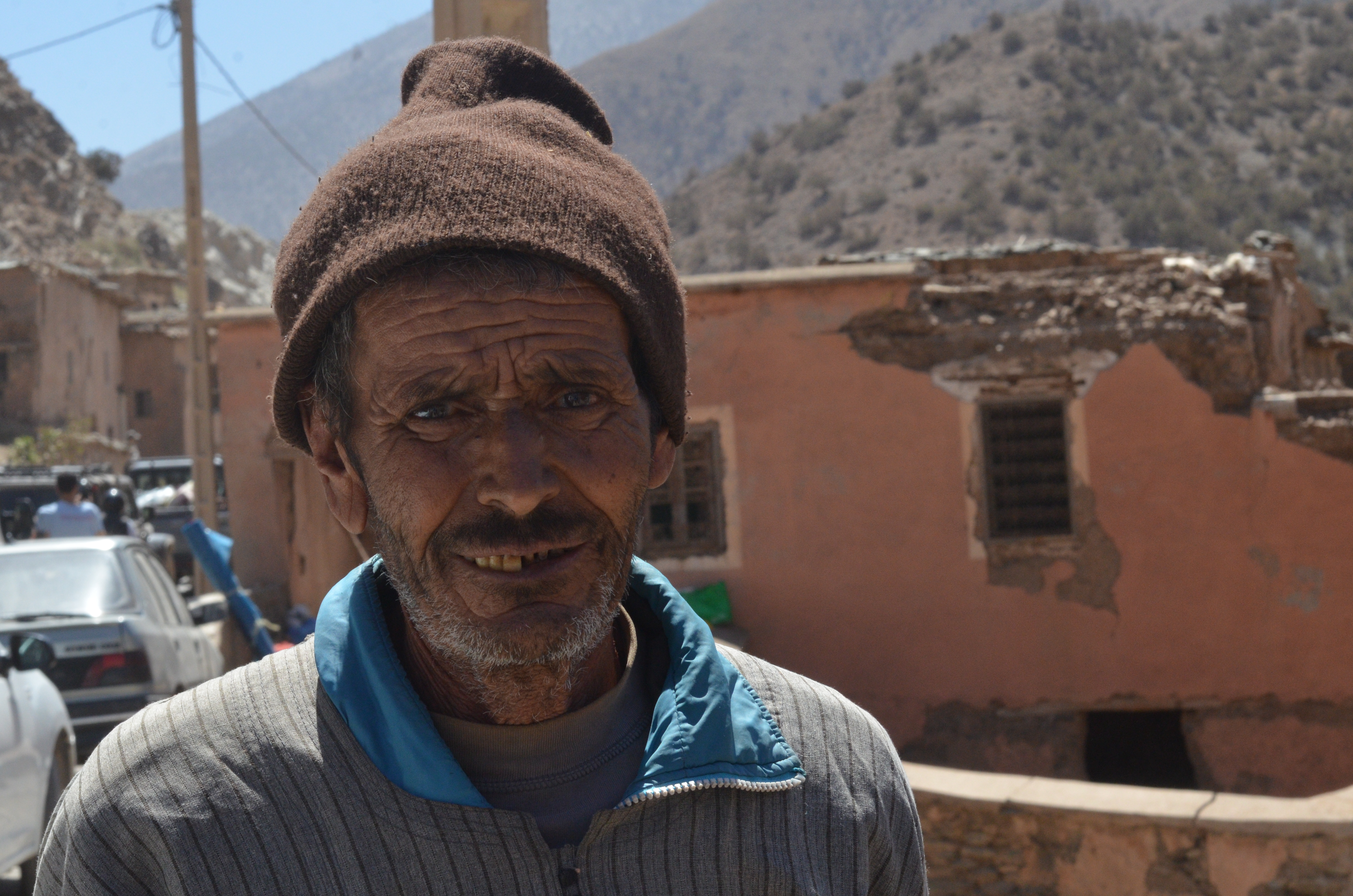 A man in a brown beanie hat stands in front of a building that is missing pieces.