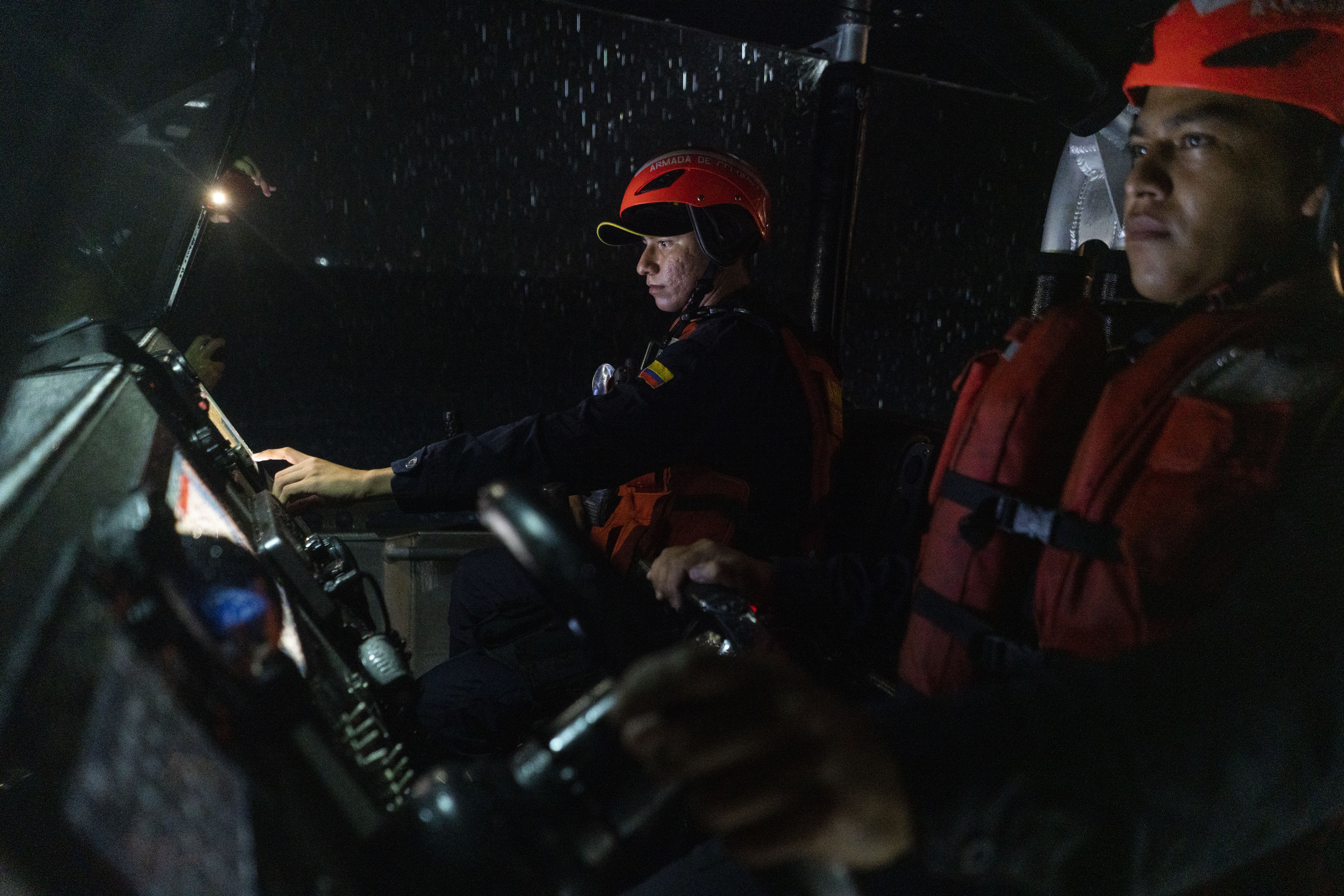 Coast Guard members, dressed in orange helmets and life vests, stand behind the controls of their boat during a nighttime patrol.