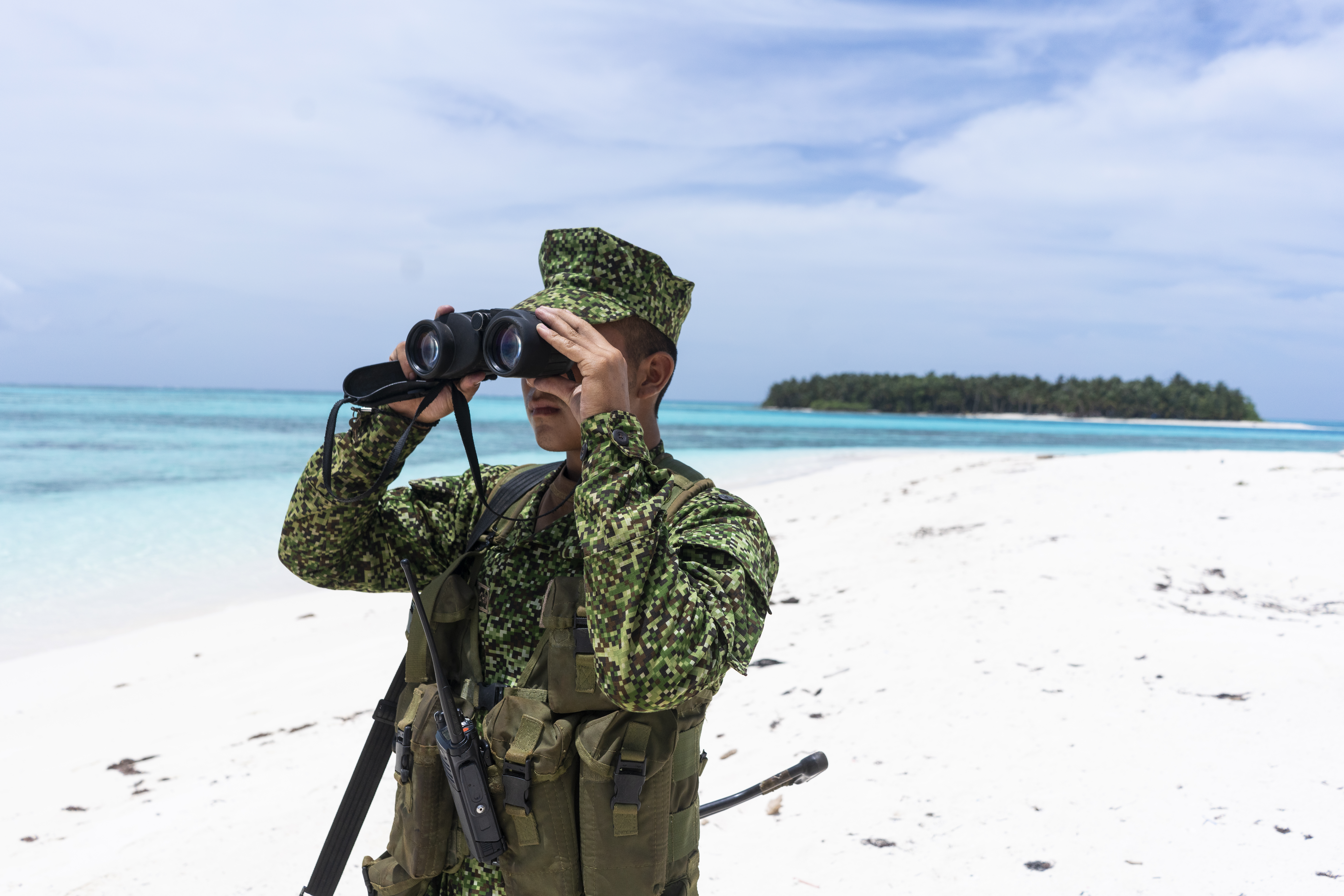 A Colombian naval officer dressed in olive fatigues looks across a white-sand beach with binoculars.