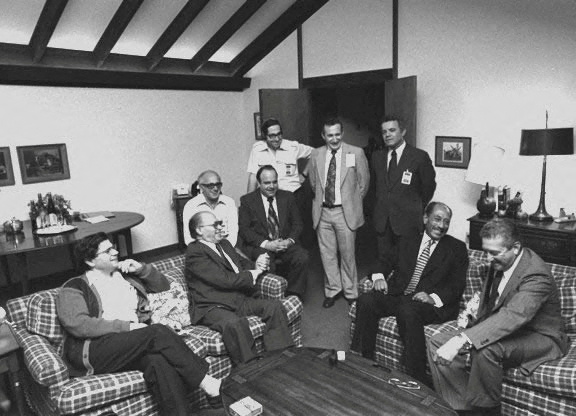 A group of men in suits and collared shirts stand around or sit on plaid couches in a room at Camp David.