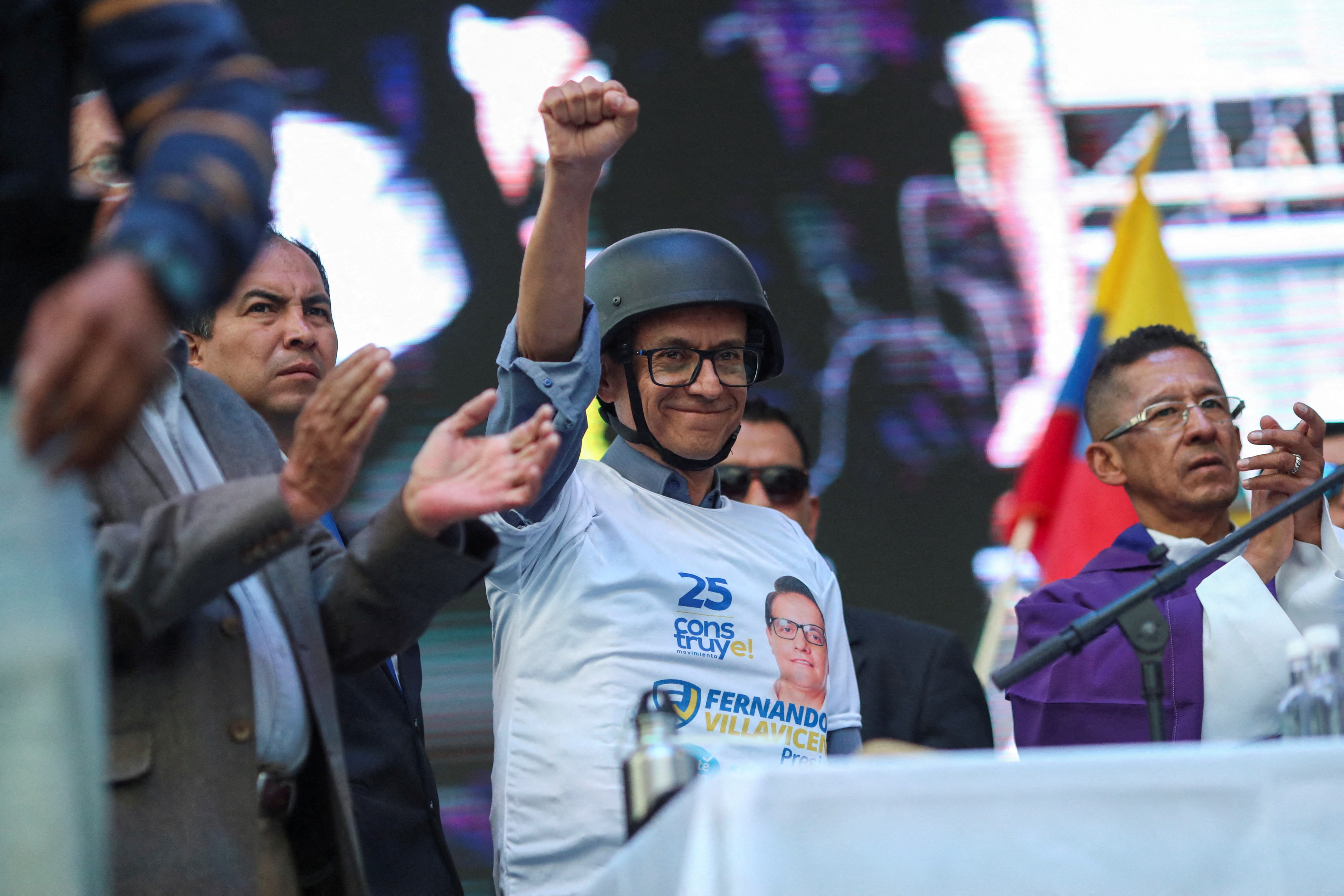 A man in a helmet and a white T-shirt raises a fist triumphantly from a podium while smiling.