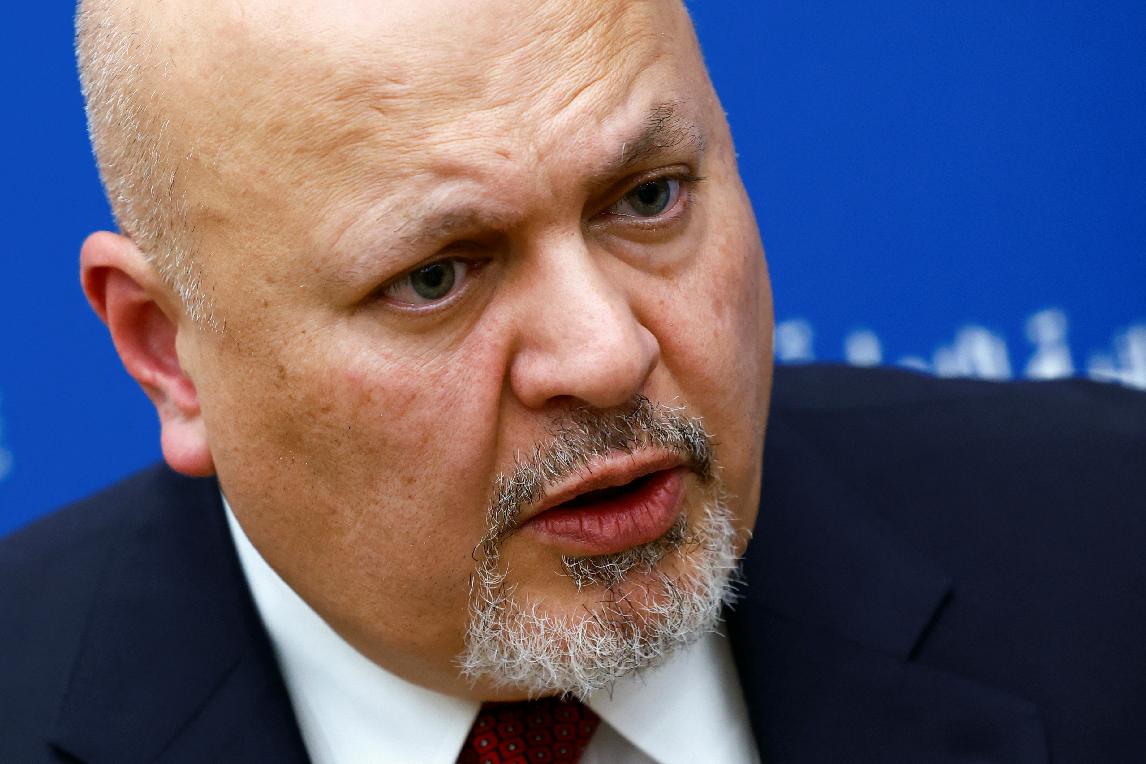 a close up of the face of a man with a grey beard wearing a suit with a blue background