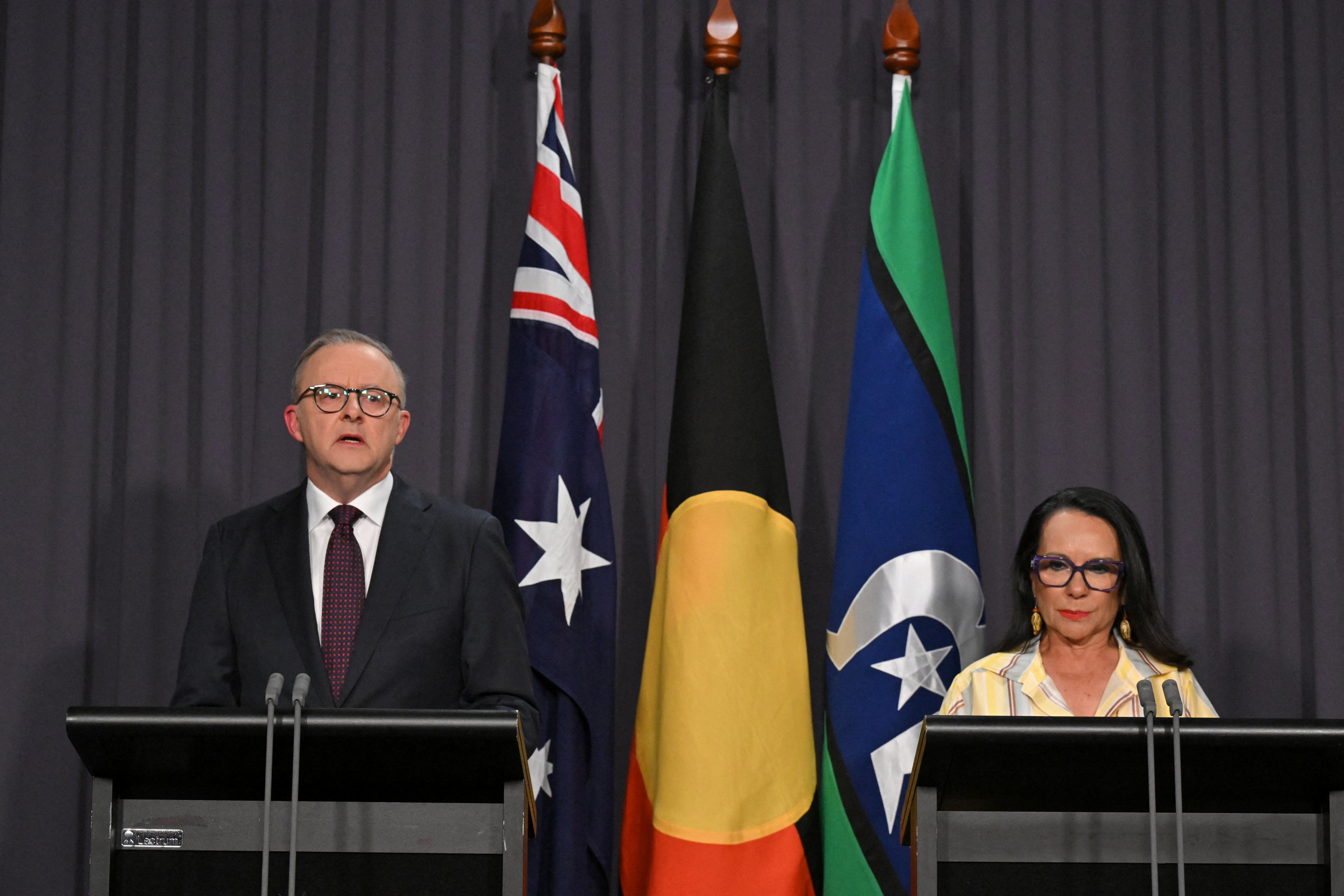 A man in a black suit and tie stands next to a woman in yellow both standing in front of podiums with 3 flags in the background Australian, Torres Strait and Aboriginal