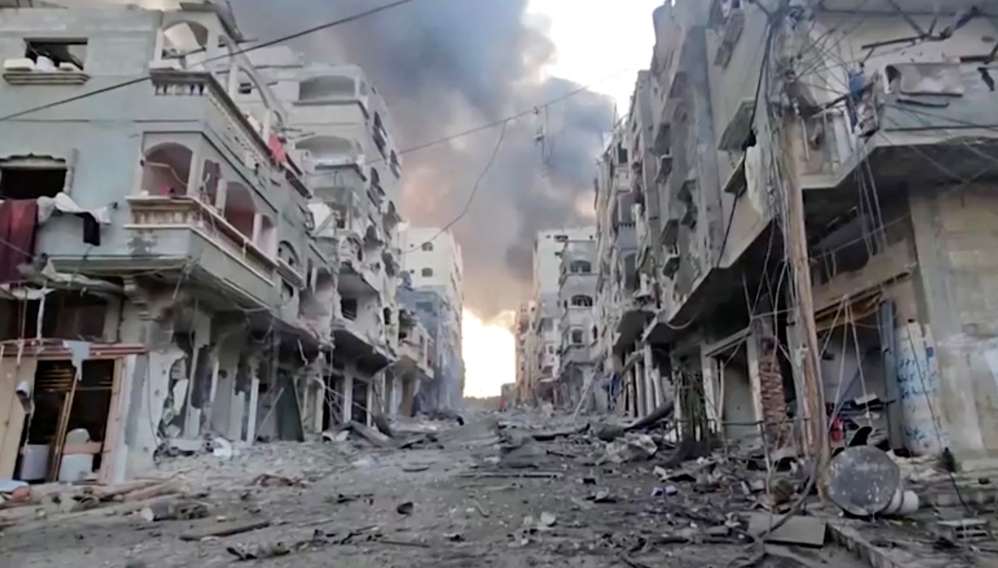 A view down the centre of a bombed-out street, as clouds of smoke hang in the air and buildings are grey with dust and rubble.