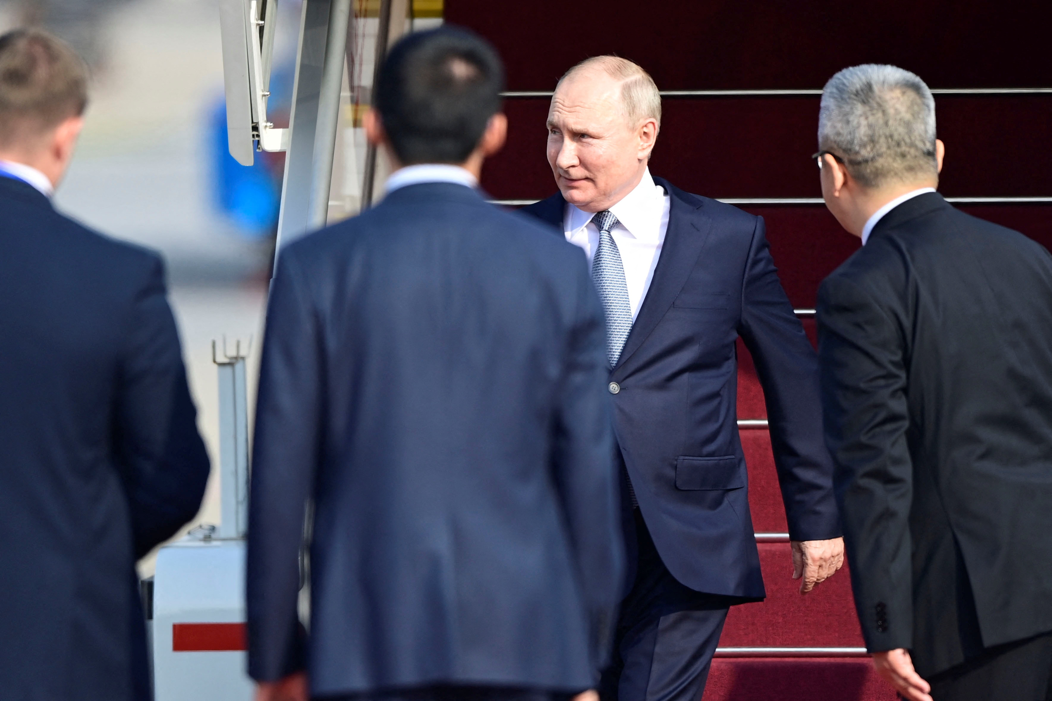 a man in a suit steps off airplane steps with red carpet as men wait to greet him