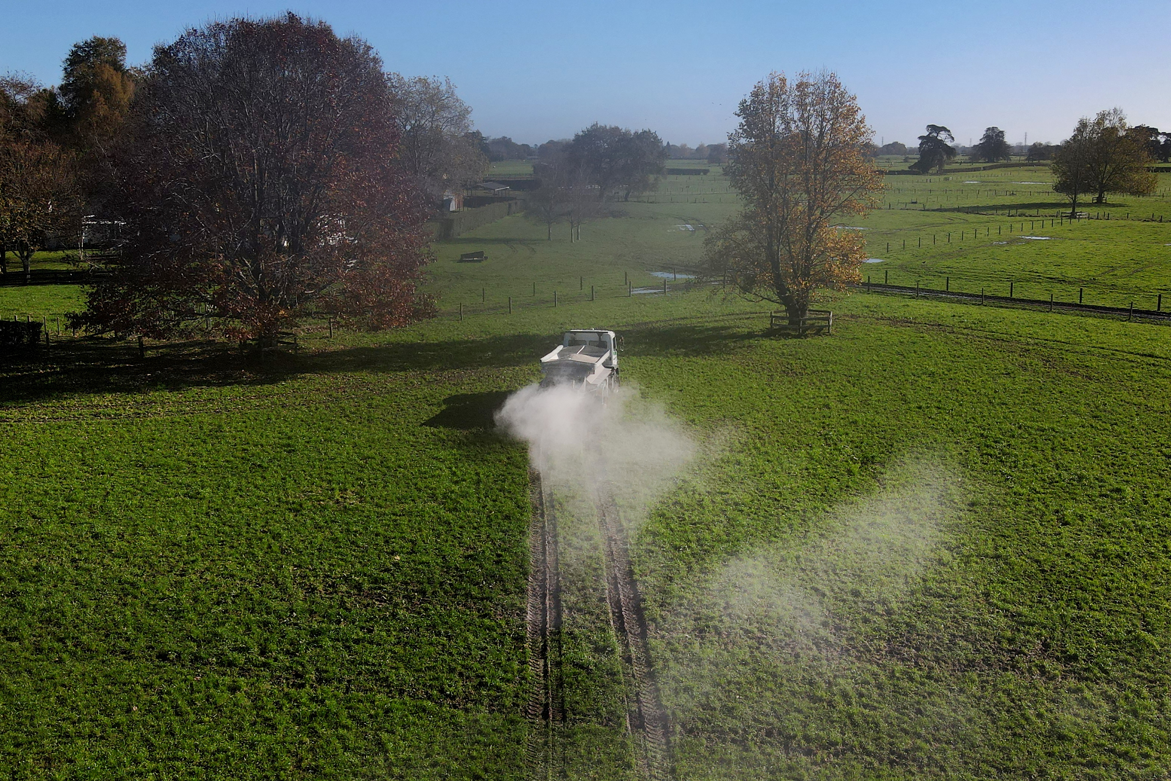 Clouds of fertiliser trail behind a white truck in a green field with scattered trees in the distance. 