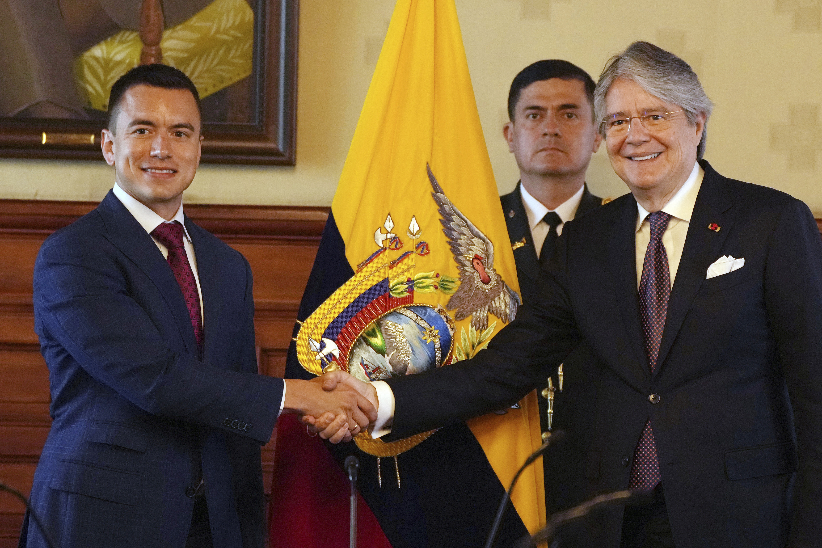 Daniel Noboa shakes hands with Guillermo Lasso. Behind them is an Ecuador flag and a man standing against a wall in a suit.