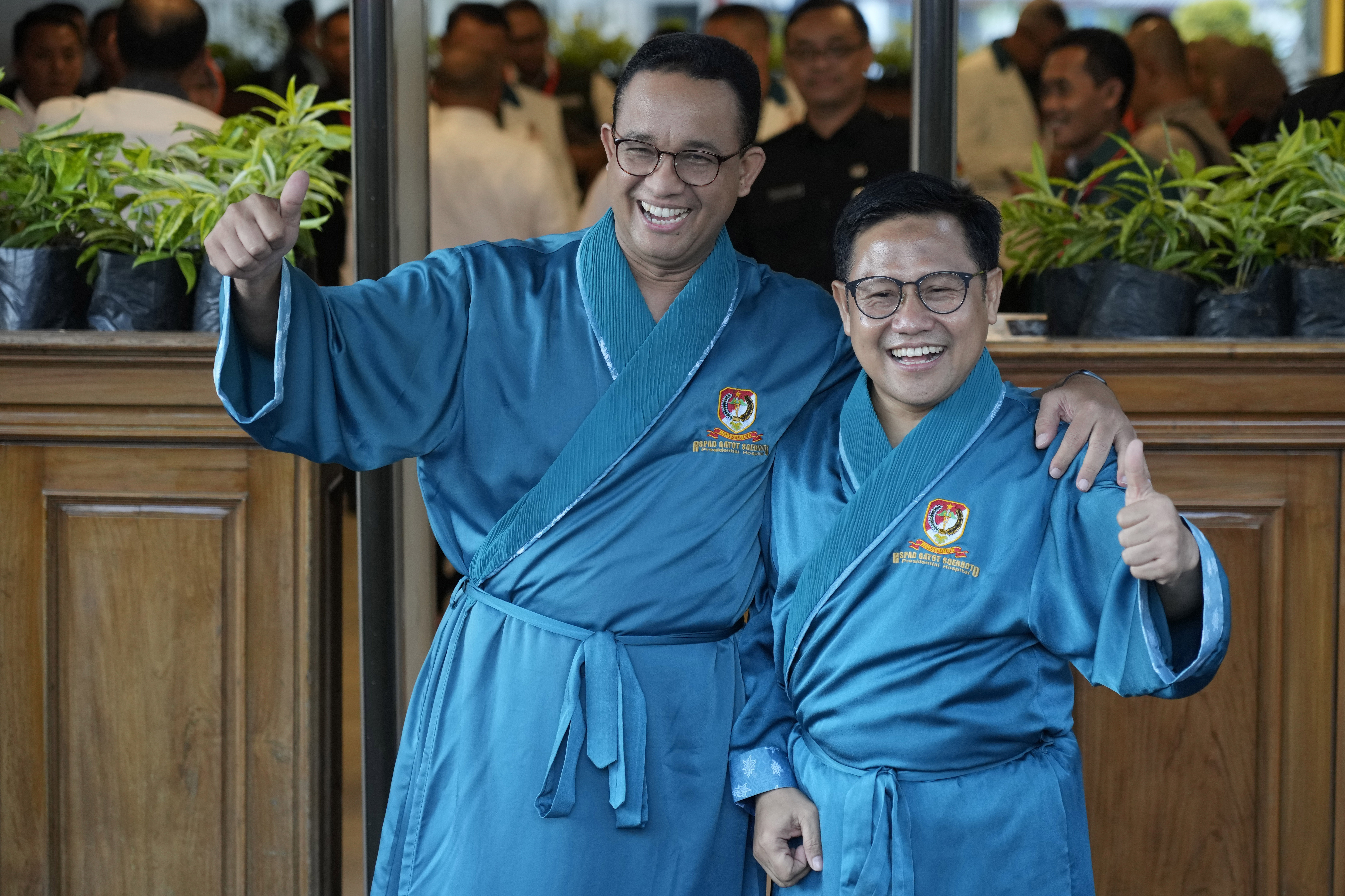 Anies Baswedan, left, and his running mate Muhaimin Iskandar smile at reporters and give a thumbs-up. They are wearing blue robes ahead of a medical check-up required of every candidate