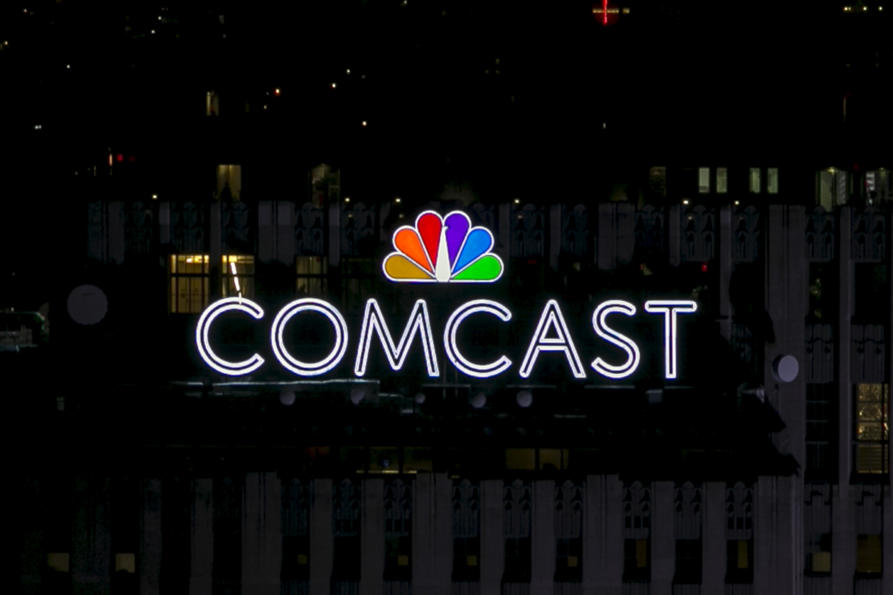 A sign featuring the COMCAST NBC logos glows in the dark atop a building.