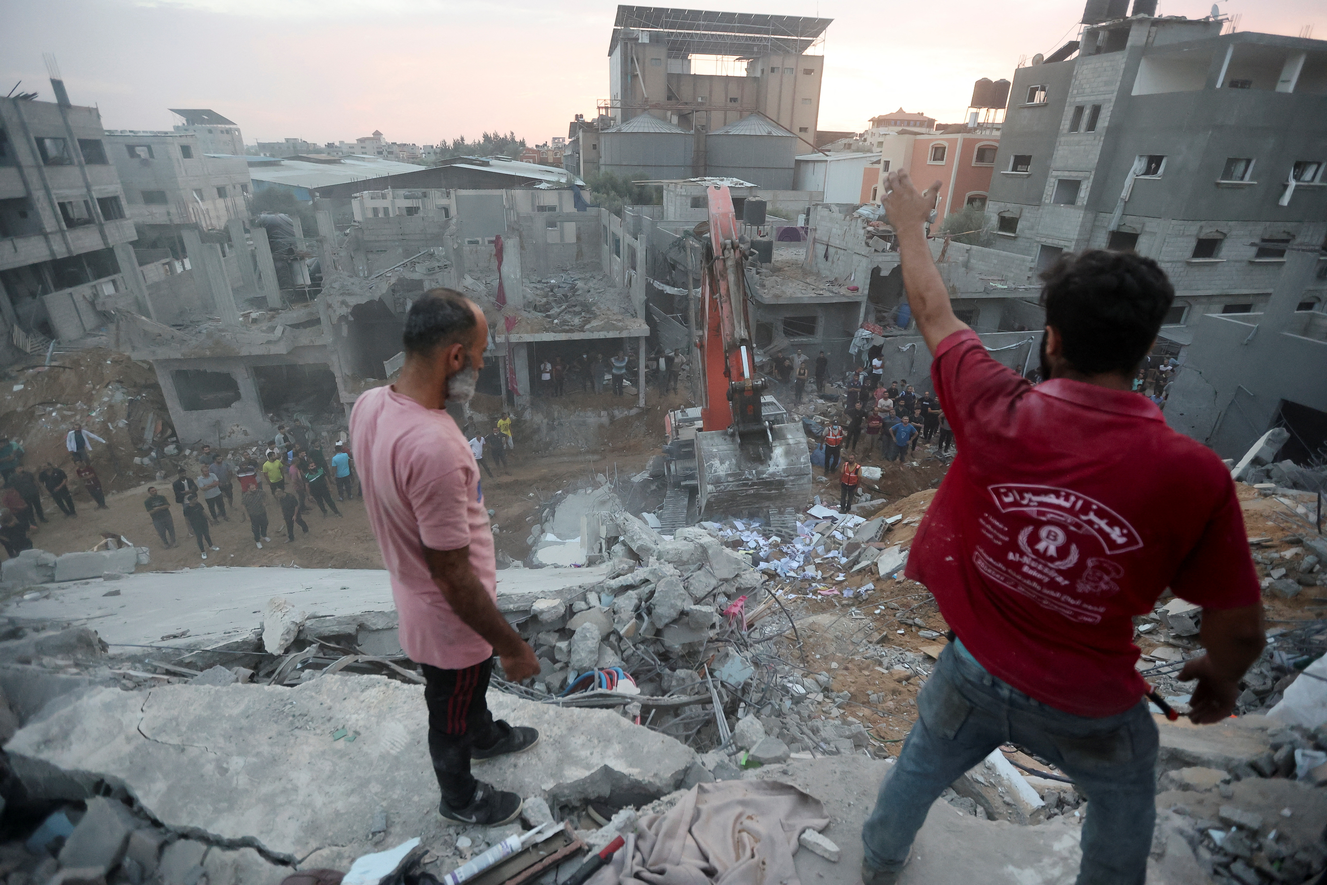 Palestinians searching for survivors in the rubble of a collapsed building. Two men are standing on the debris. An excavator is below. There are more damaged building behind