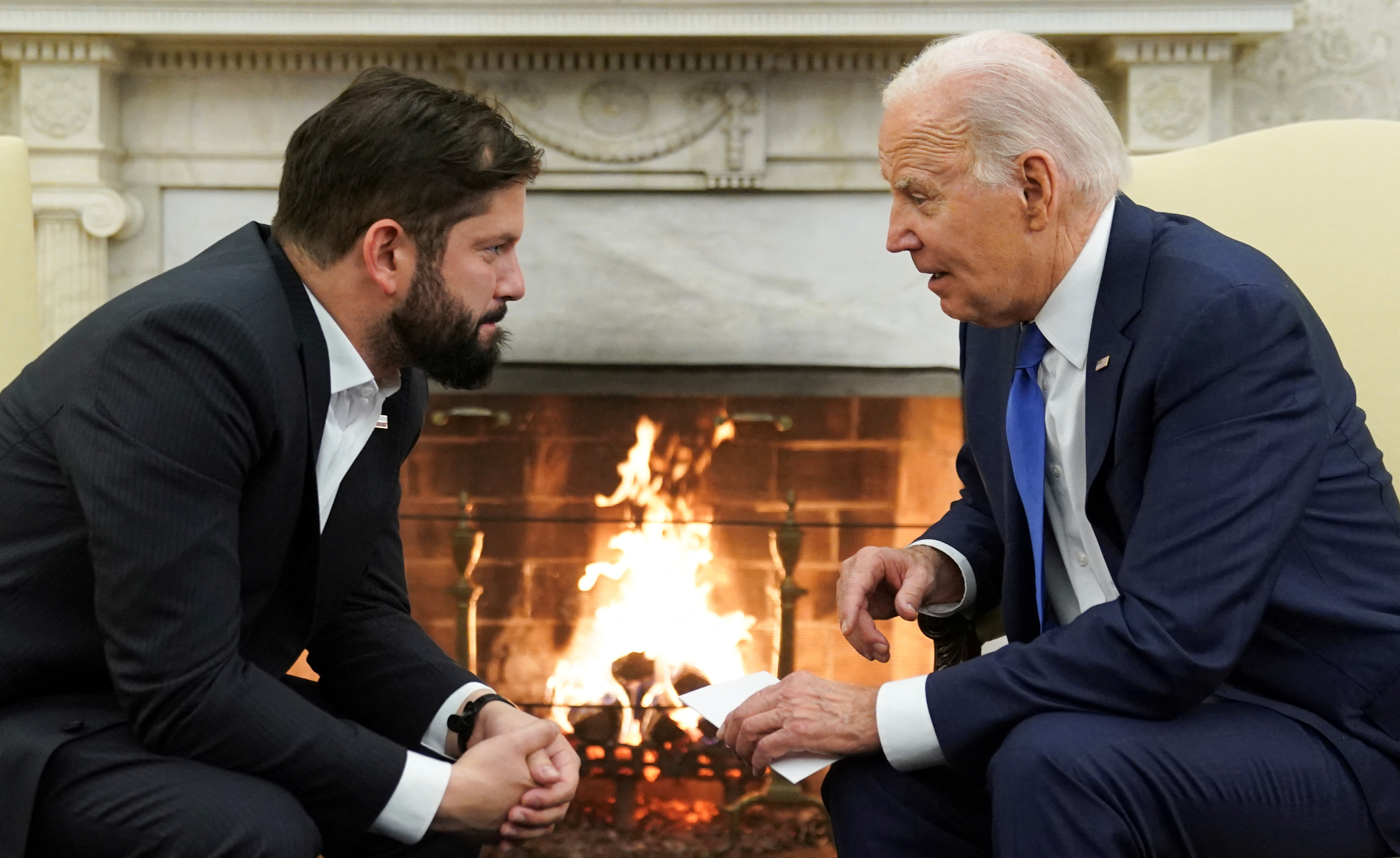 Gabriel Boric and Joe Biden face each other and talk in the Oval Office, a roaring fire in the fireplace behind them.