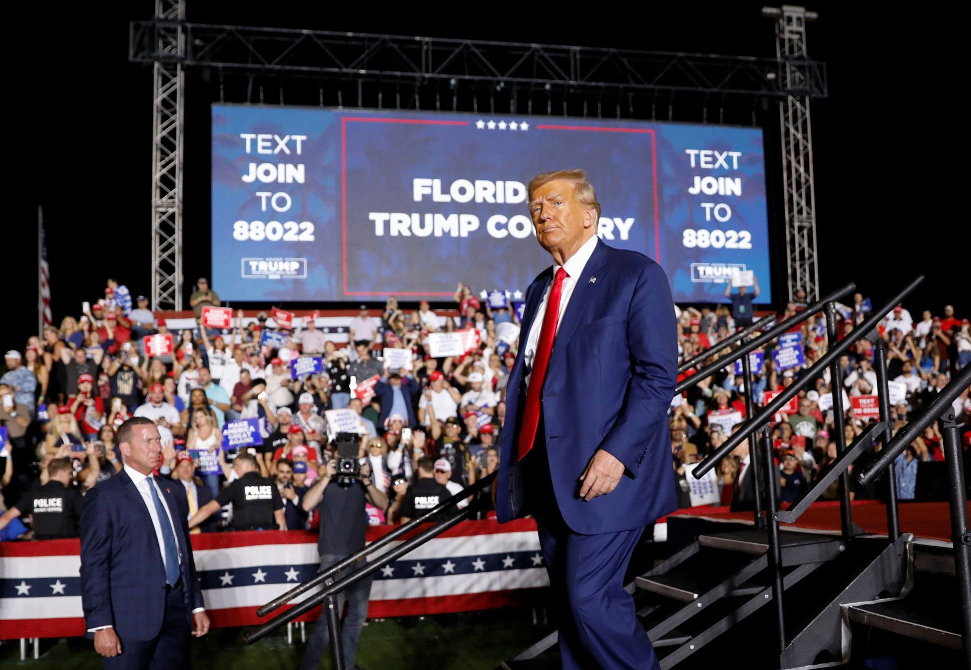Trump, in a blue suit and red tie, walks down a metal staircase in front of a crowd of supporters, who cheer in front of a sign that reads: "Florida is Trump country"