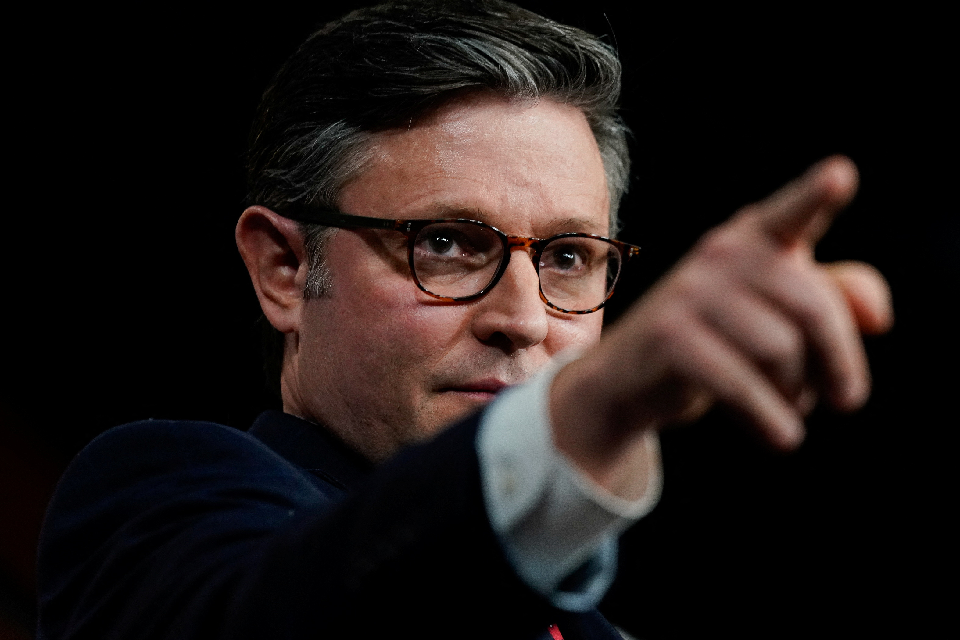 Mike Johnson — a man wearing glasses and a suit and tie — points in front of himself.