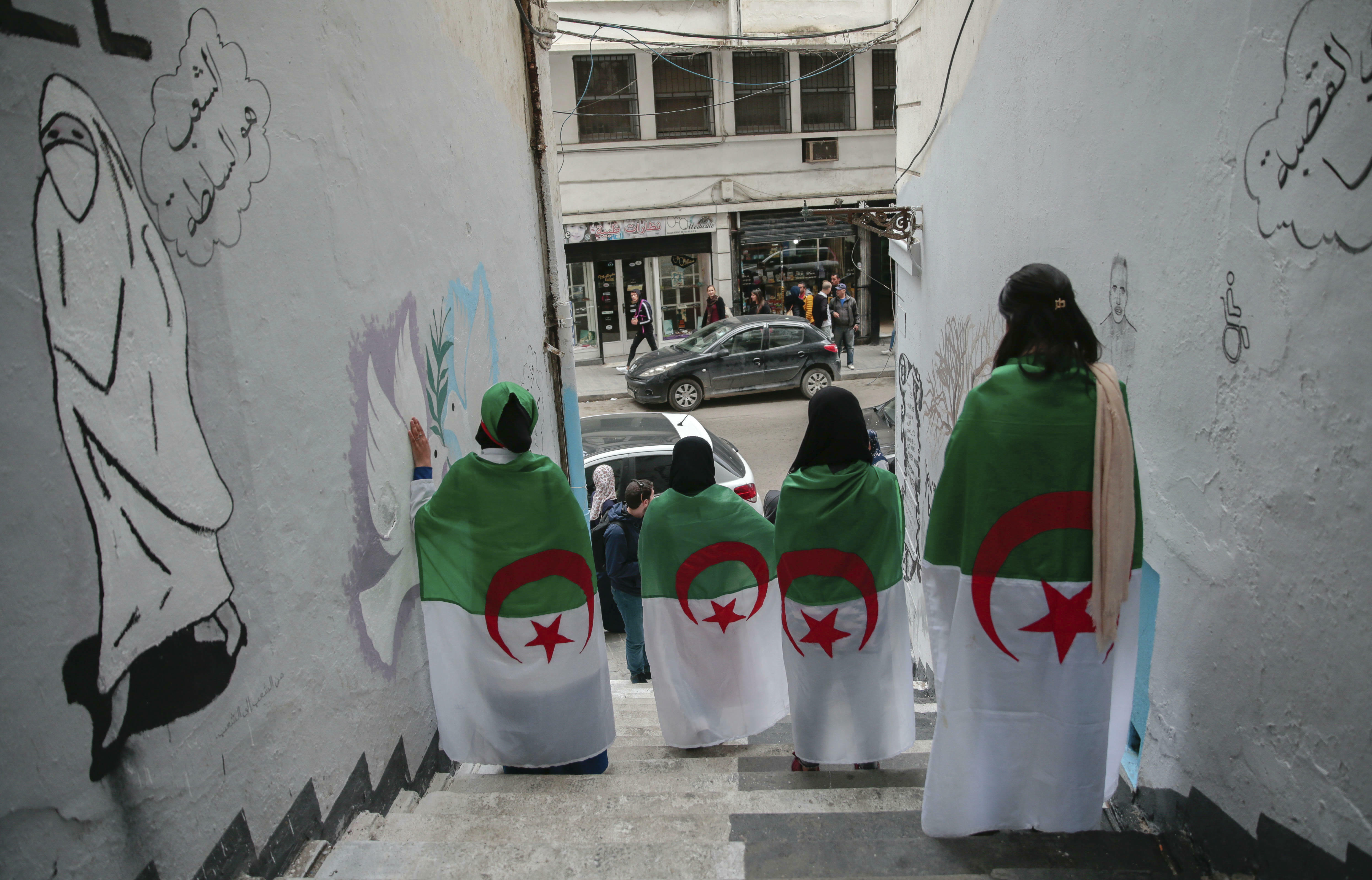 young Algerian women wrapped in Algerian flags pose next to street art