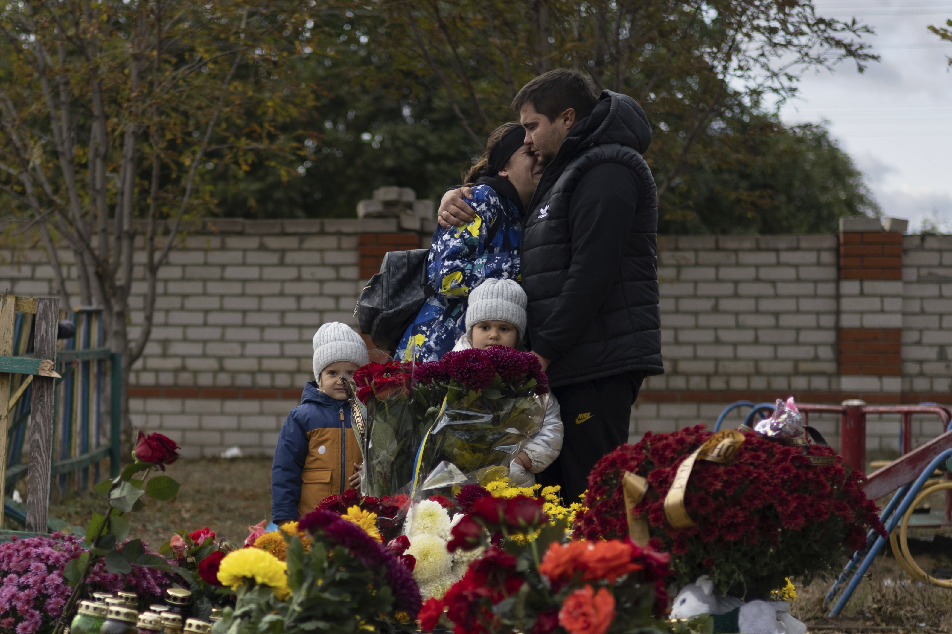 A family visit a memorial for those killed in Hroza. There are lots of flowers. The parents are hugging each other