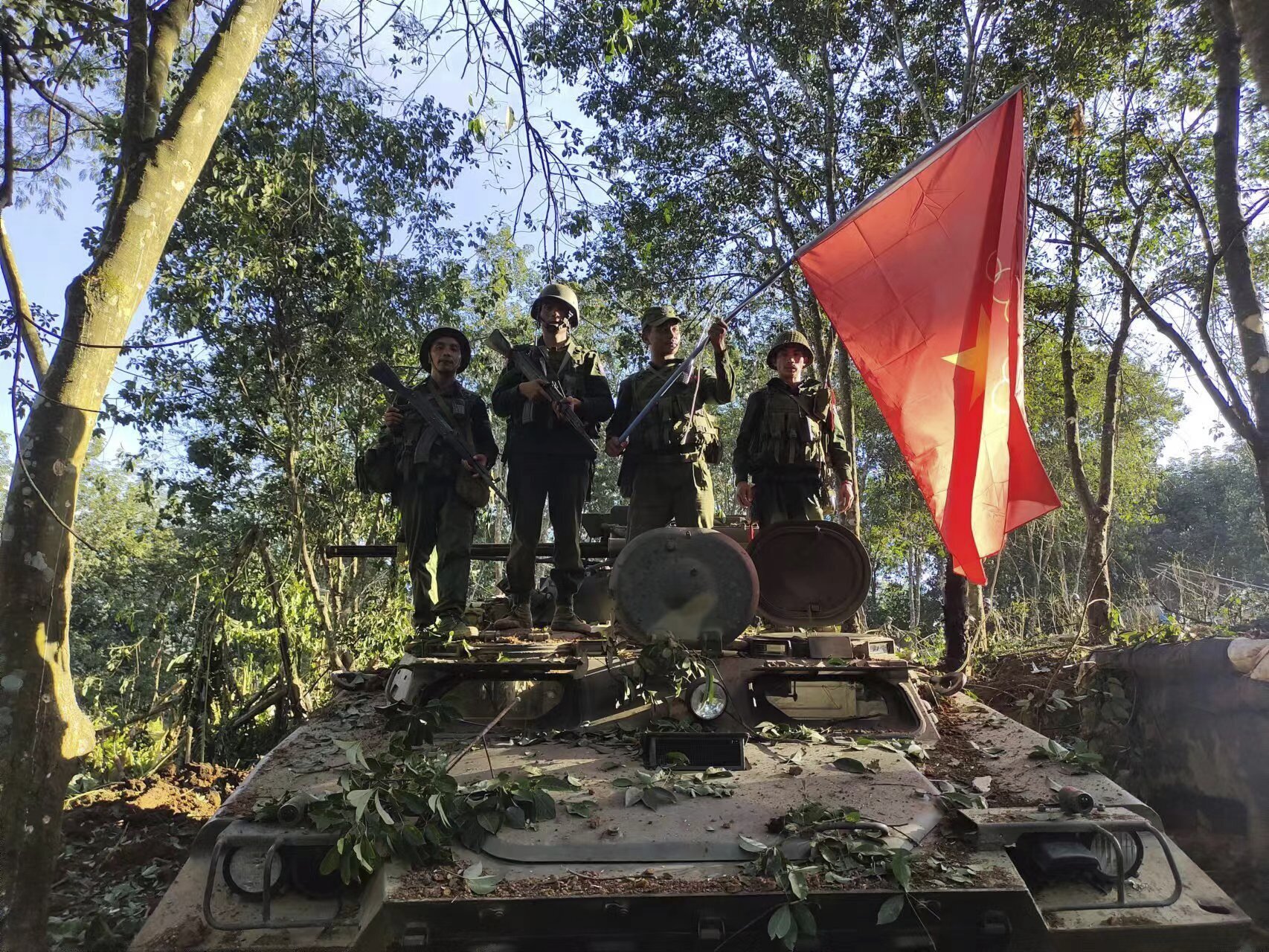 Soldiers from the Myanmar National Democratic Alliance Army standing on top of a capture Myanmar military tank. They have hung their flag on the tank. They are surrounded by trees.