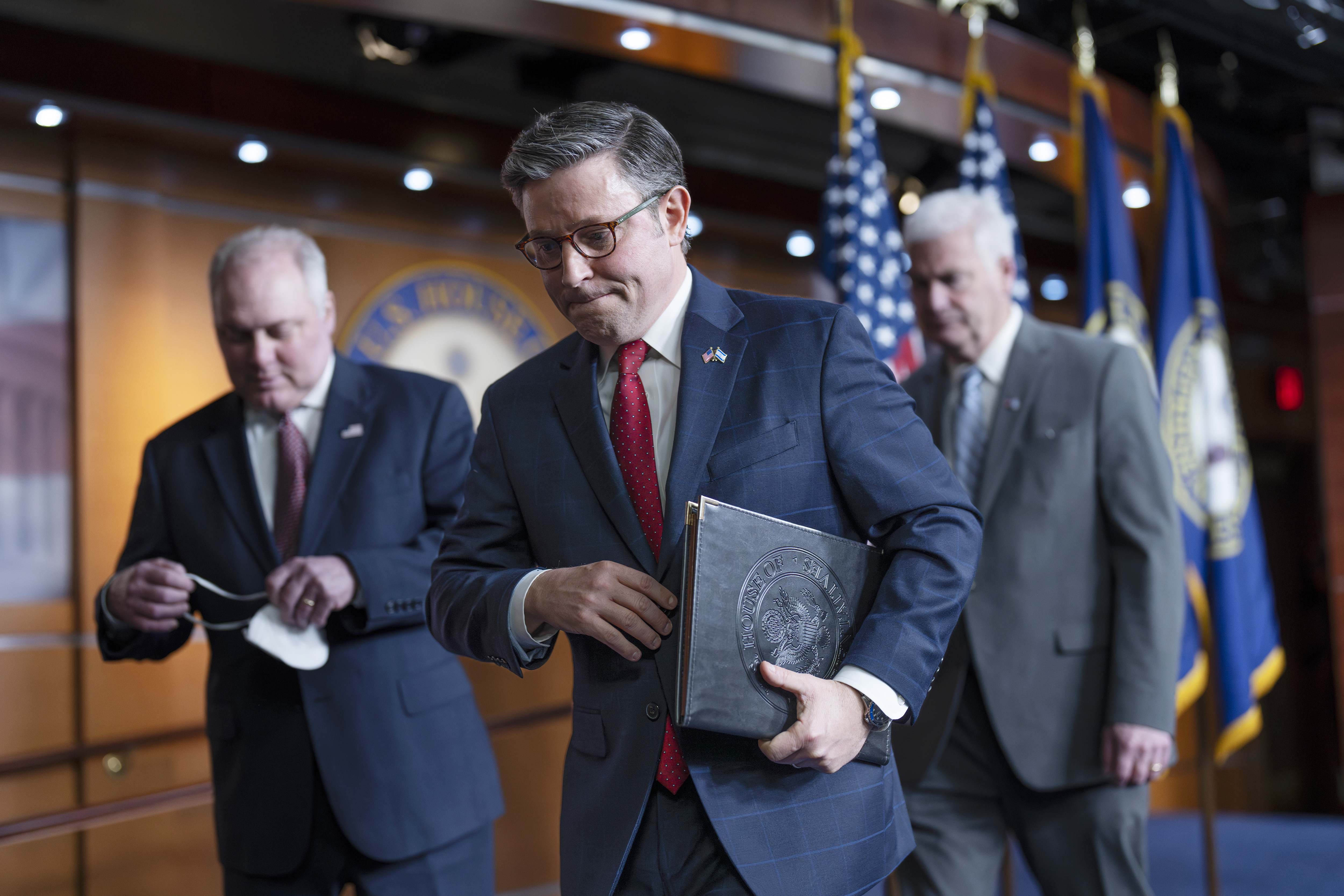 Mike Johnson, surrounded by Steve Scalise and Tom Emmer, walk away from a podium in a wood-paneled room in the Capitol. The three men wear suits, and Johnson carries a folder of documents under his arm.