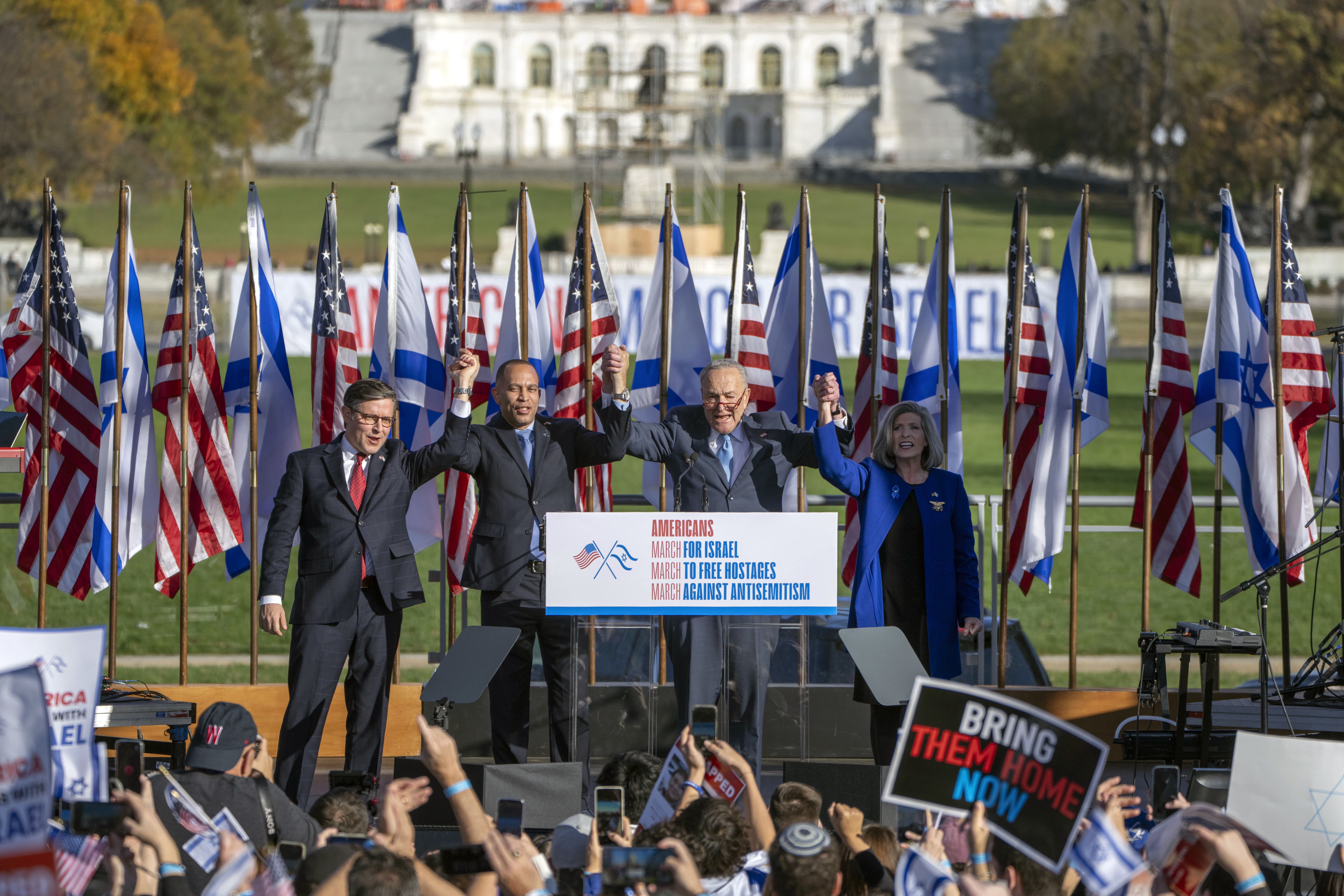 Speaker of the House Mike Johnson of La., left, House Minority Leader Hakeem Jeffries of N.Y., Senate Majority Leader Chuck Schumer of N.Y., and Sen. Joni Ernst, R-Iowa, right, join hands at the March for Israel. There are banners behind them and people in front
