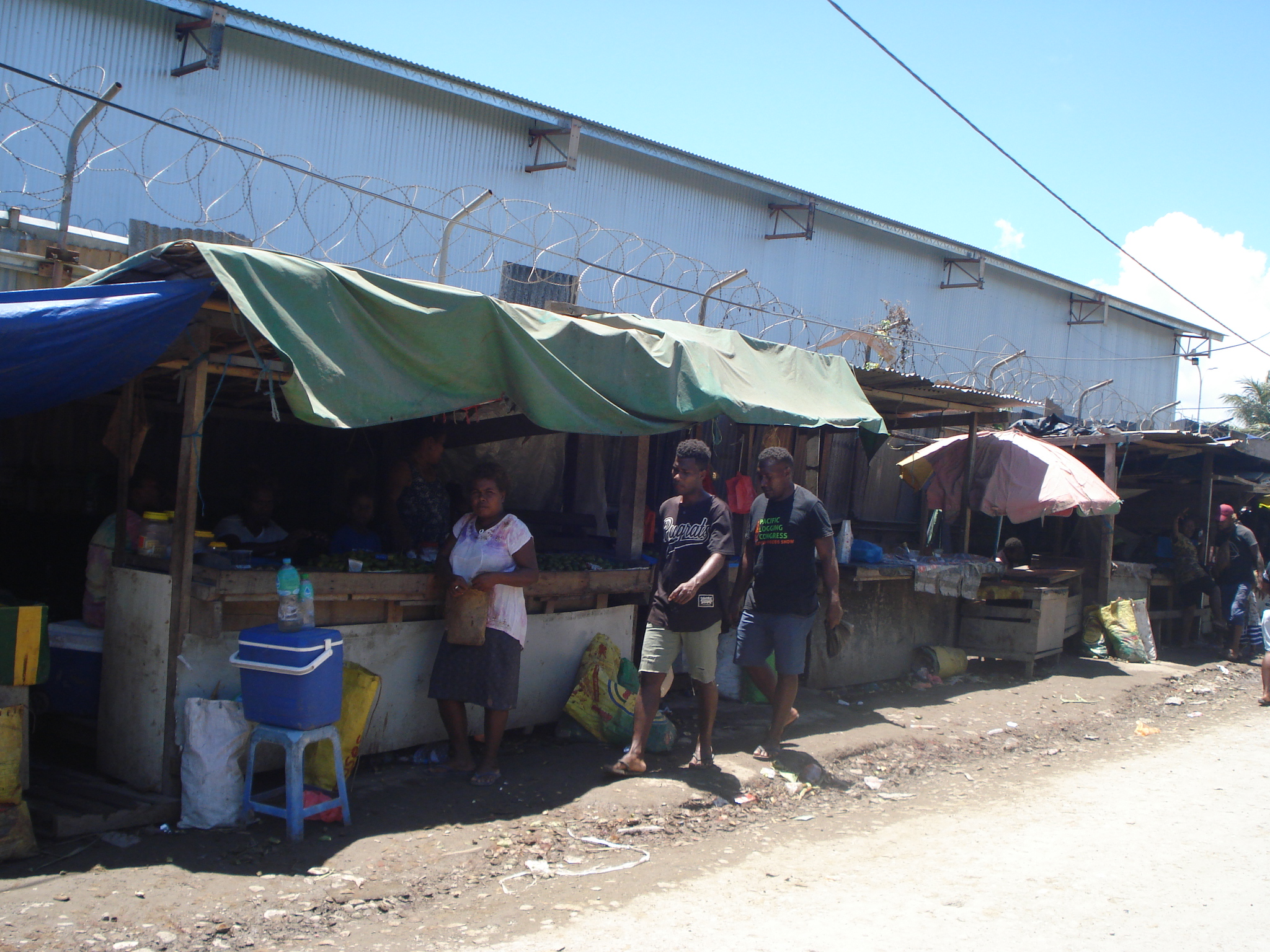 Stalls along the street in the Burns Creek informal settlement. The stalls are along a wall topped with bundles of barbed wire. A couple of young men are walking past. A woman is standing by a stall.