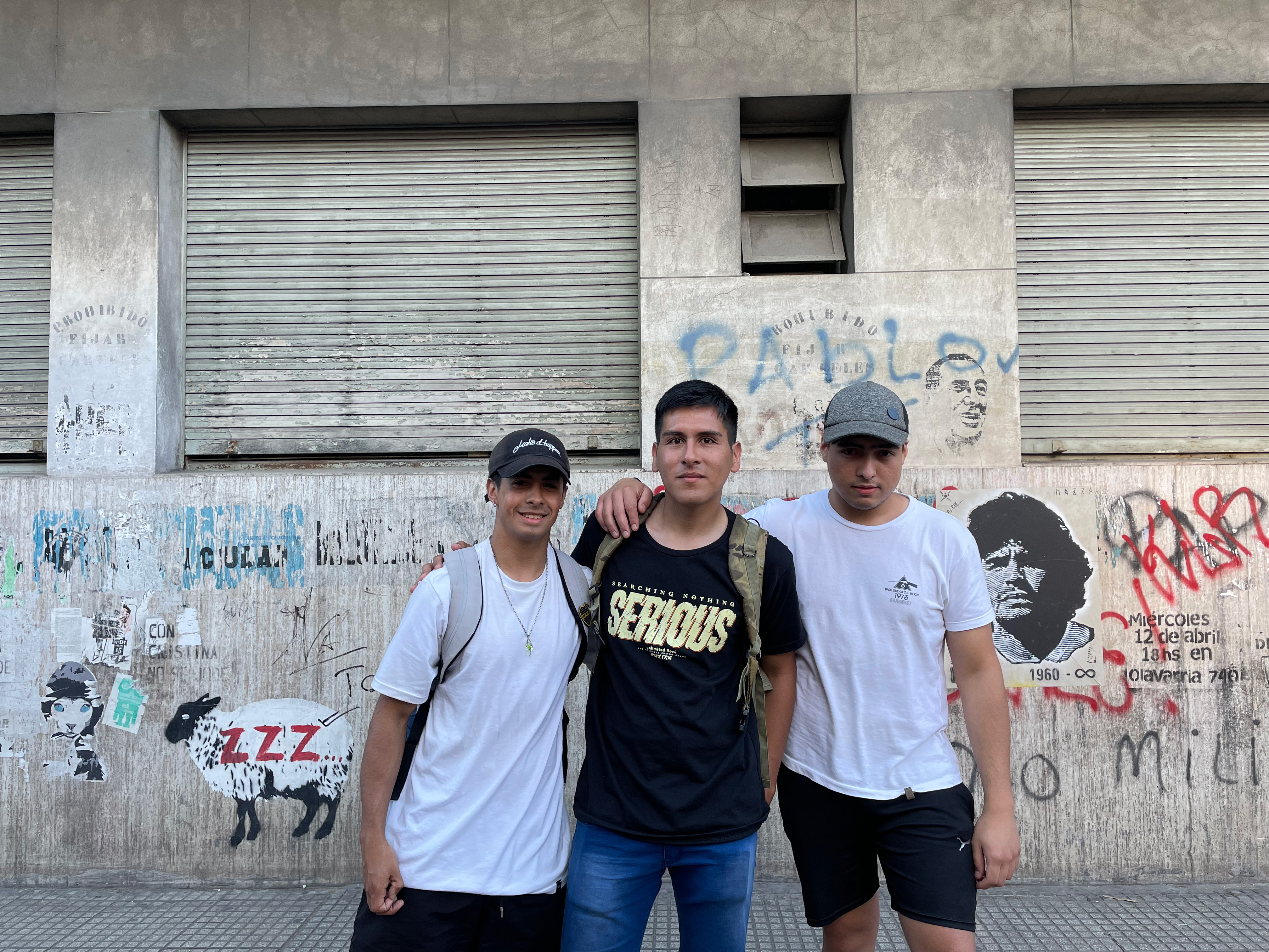 Three young men, dressed casually in T-shirts, jeans and shorts, pose for a picture together in front of a wall of graffiti.