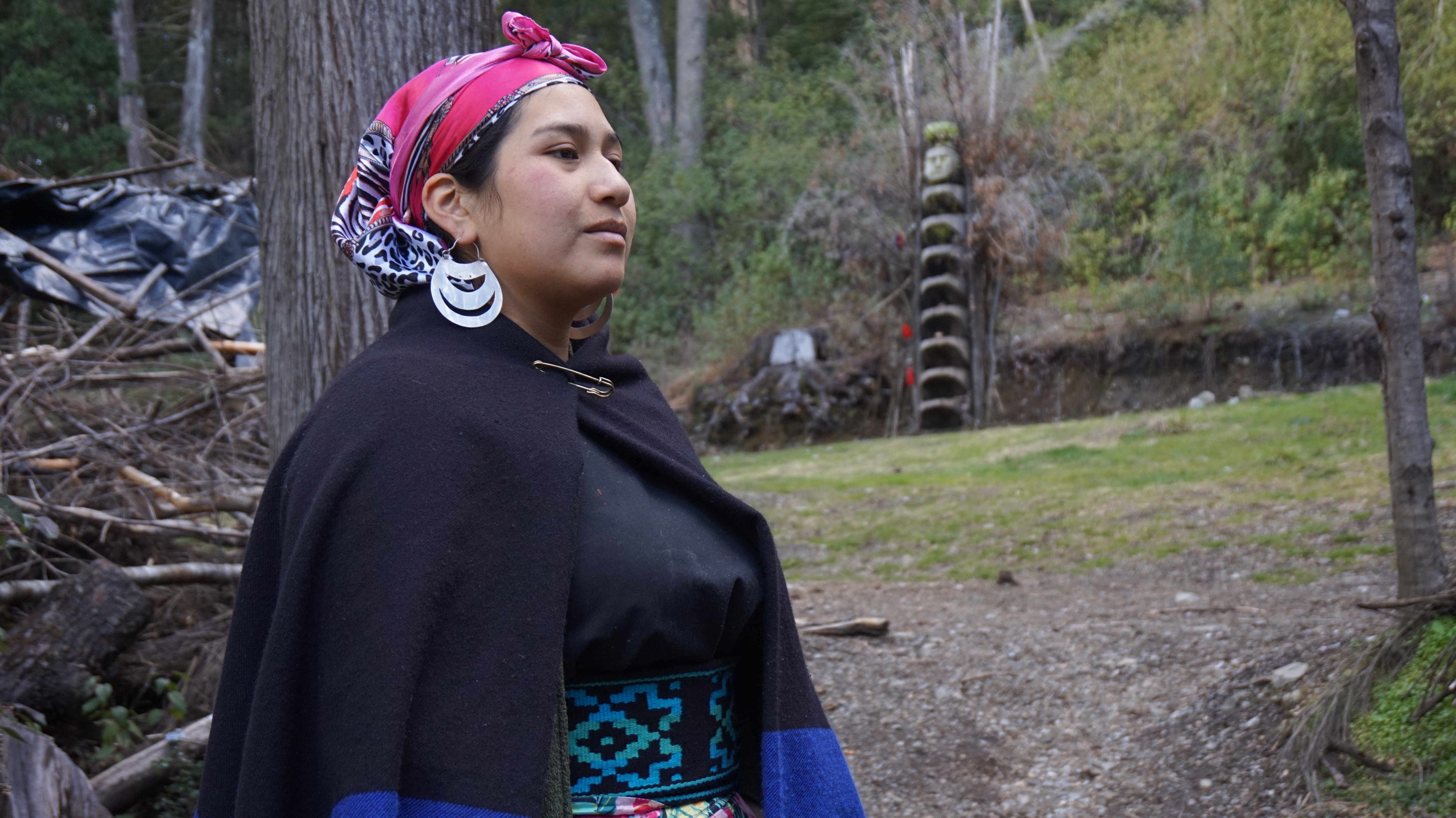 A close-up of Betiana Colhuan, a Mapuche leader. She wears a pink scarf over her hair and wears large silver earrings and a dark shawl-like garment. In the background is a green space, with a carved wooden statue signifying a rewe, a sacred space in Mapuche culture.