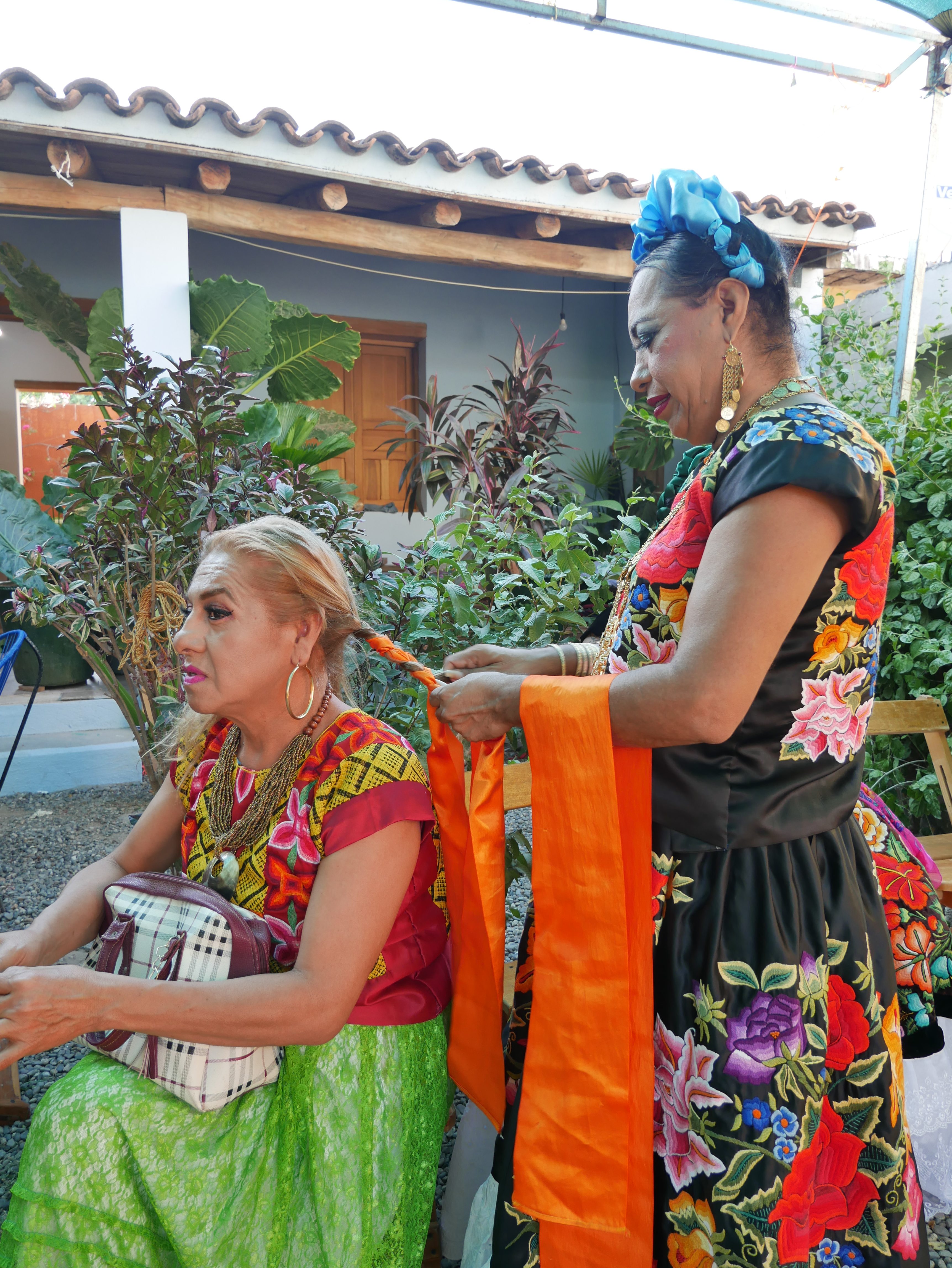 A woman in a blue headdress and a black traditional dress, decorated with flowers, braids another person's hair in the outdoor courtyard of a building, decorated with small palms.