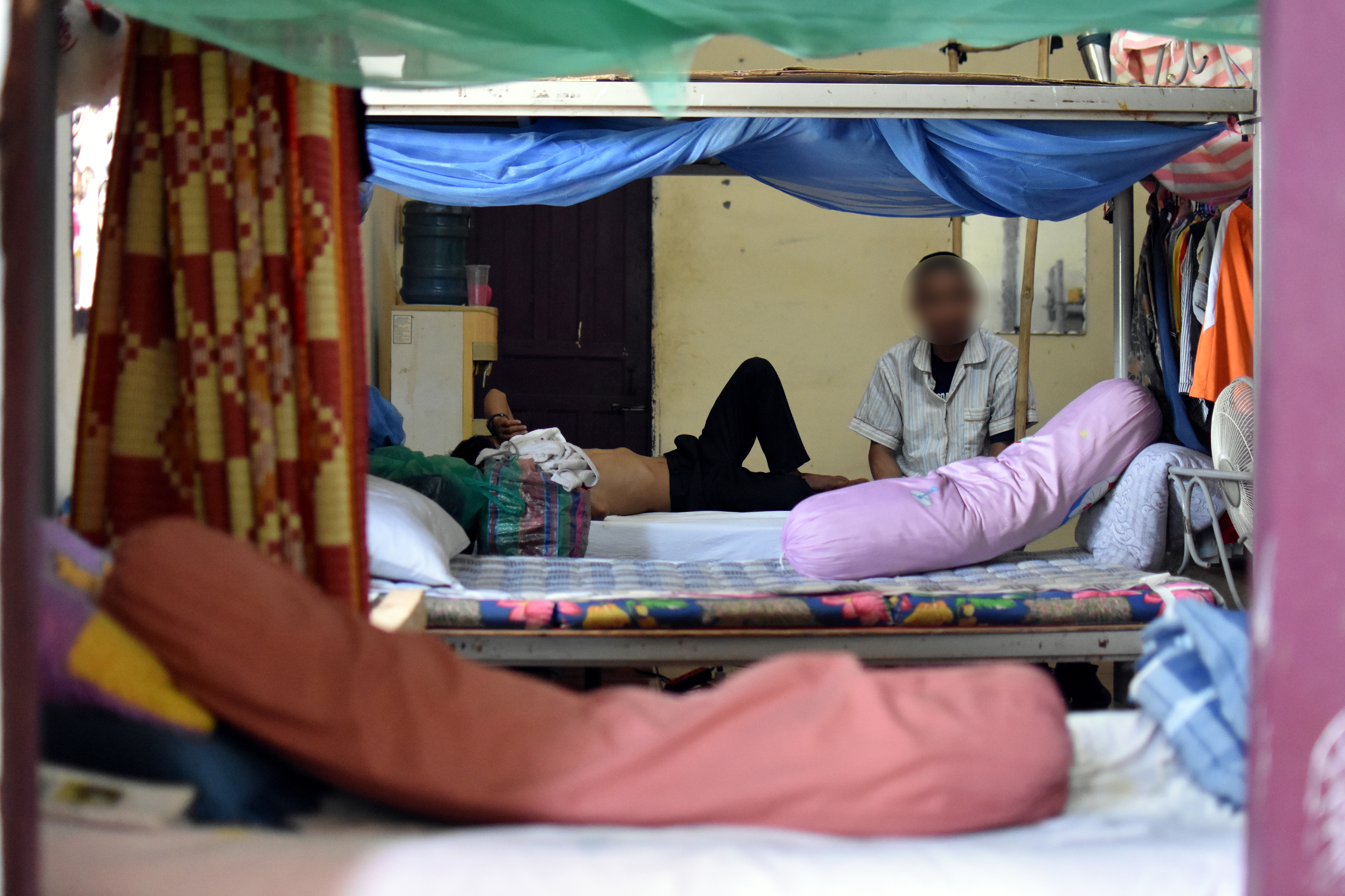 Men at a dormitory for the Transformation Center in Laos. They are sitting on beds. There are bolsters on each bed and curtains pulled to the side. 