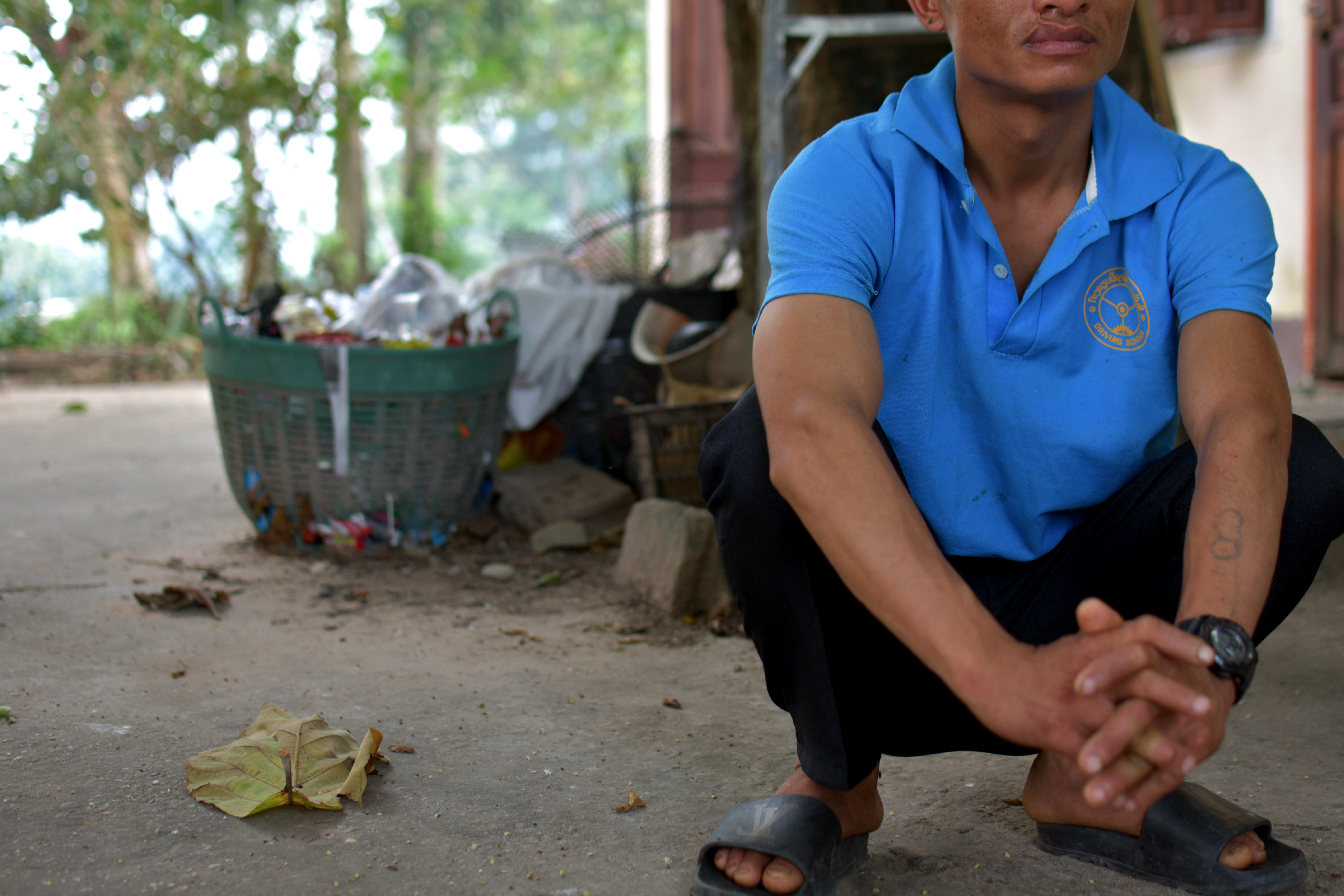 Keo at the Transformation Centre. He is wearing a blue shirt and black trousers and squatting down outside. His face is partly obscured.