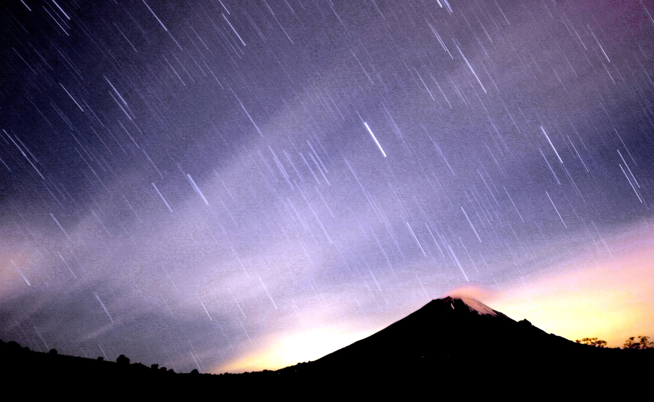 A meteor streaking past stars, light up the night sky over the Mexican volcano. Purple sky white streaks