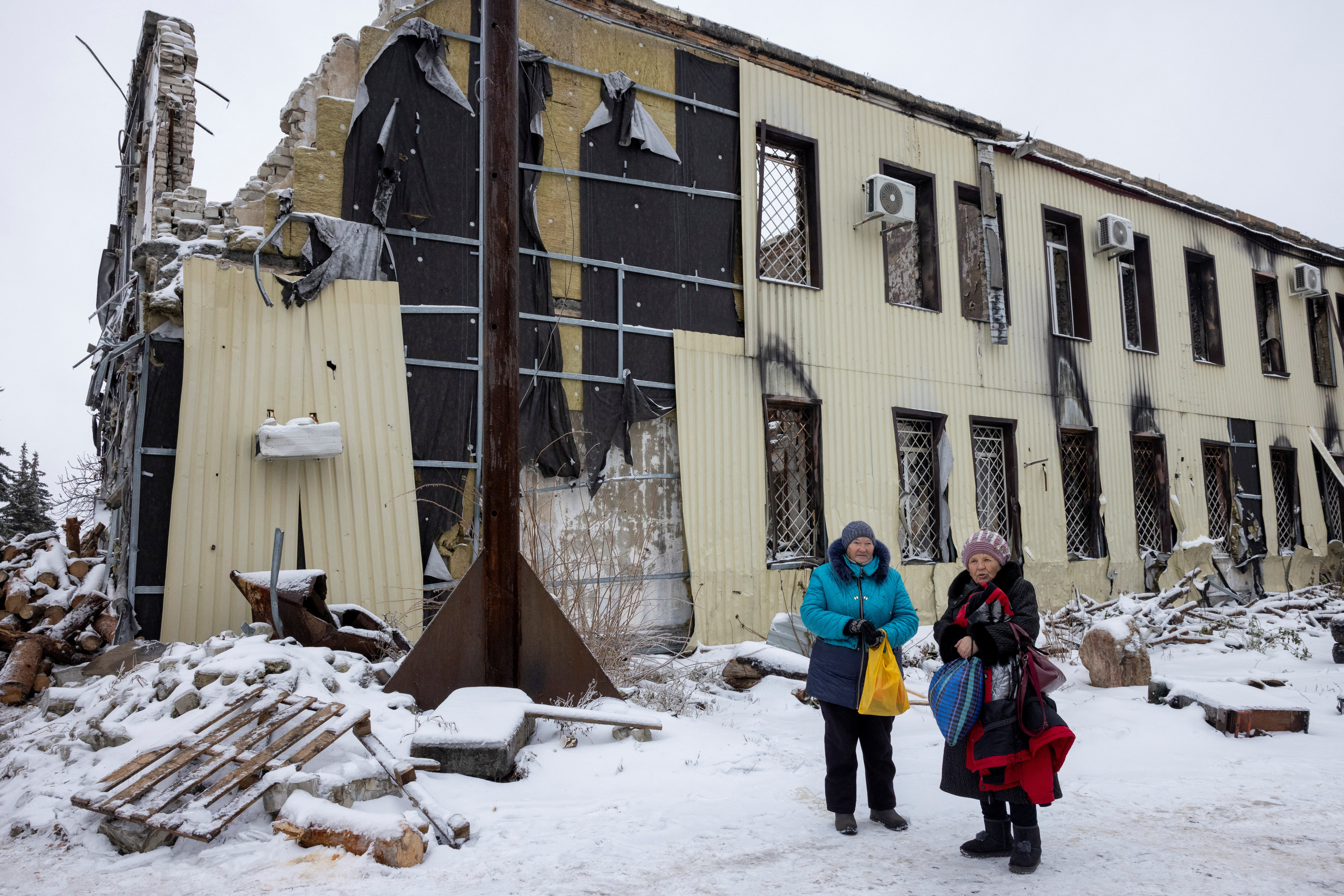 Two people collect wood in the snow next to a damaged building