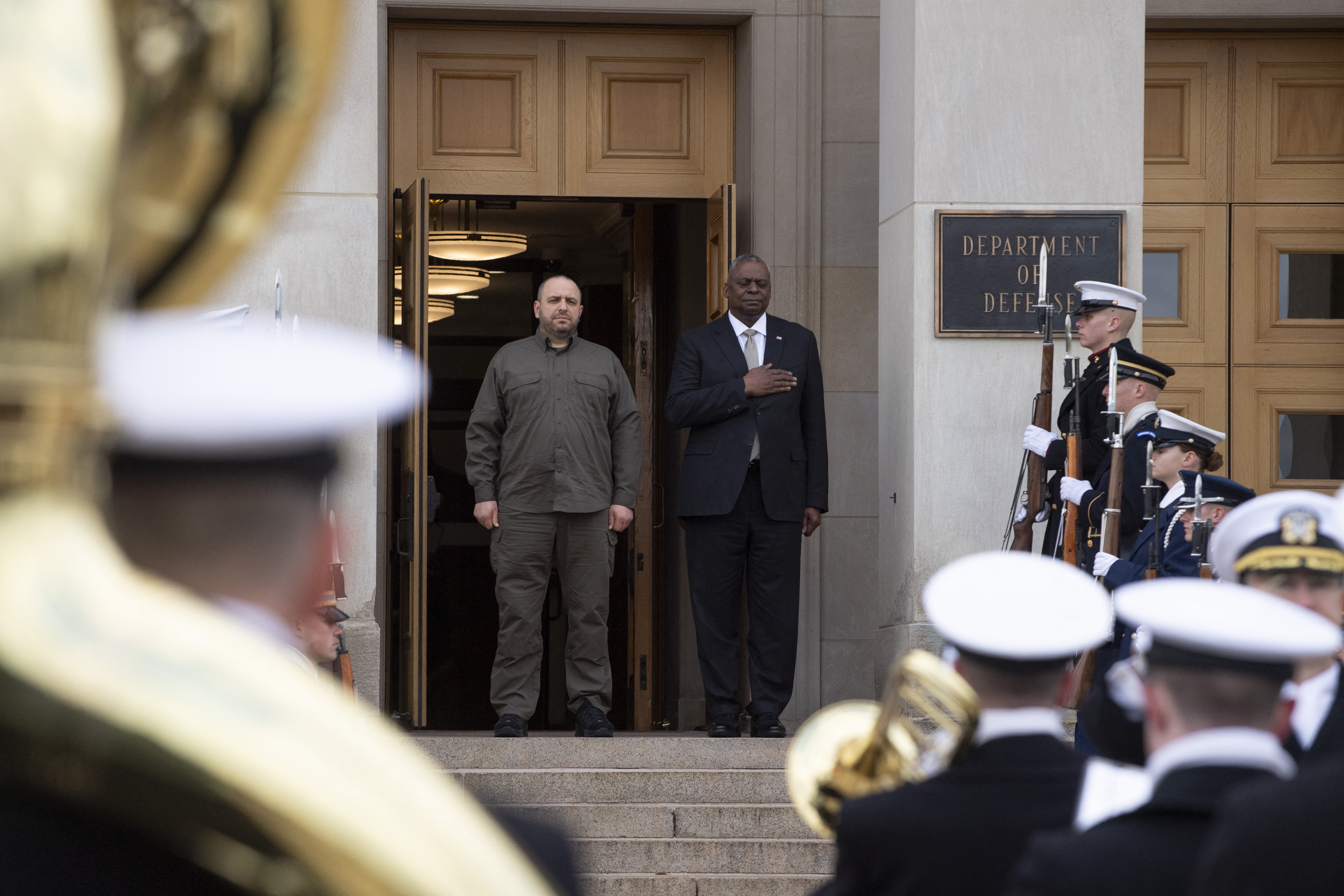 US Defense Secretary Lloyd Austin (R) and Ukrainian Defense Minister Rustem Umerov standing for the national anthems before their meeting at the Pentagon. They are standing on the steps of the building. A military band is playing in front of them.
