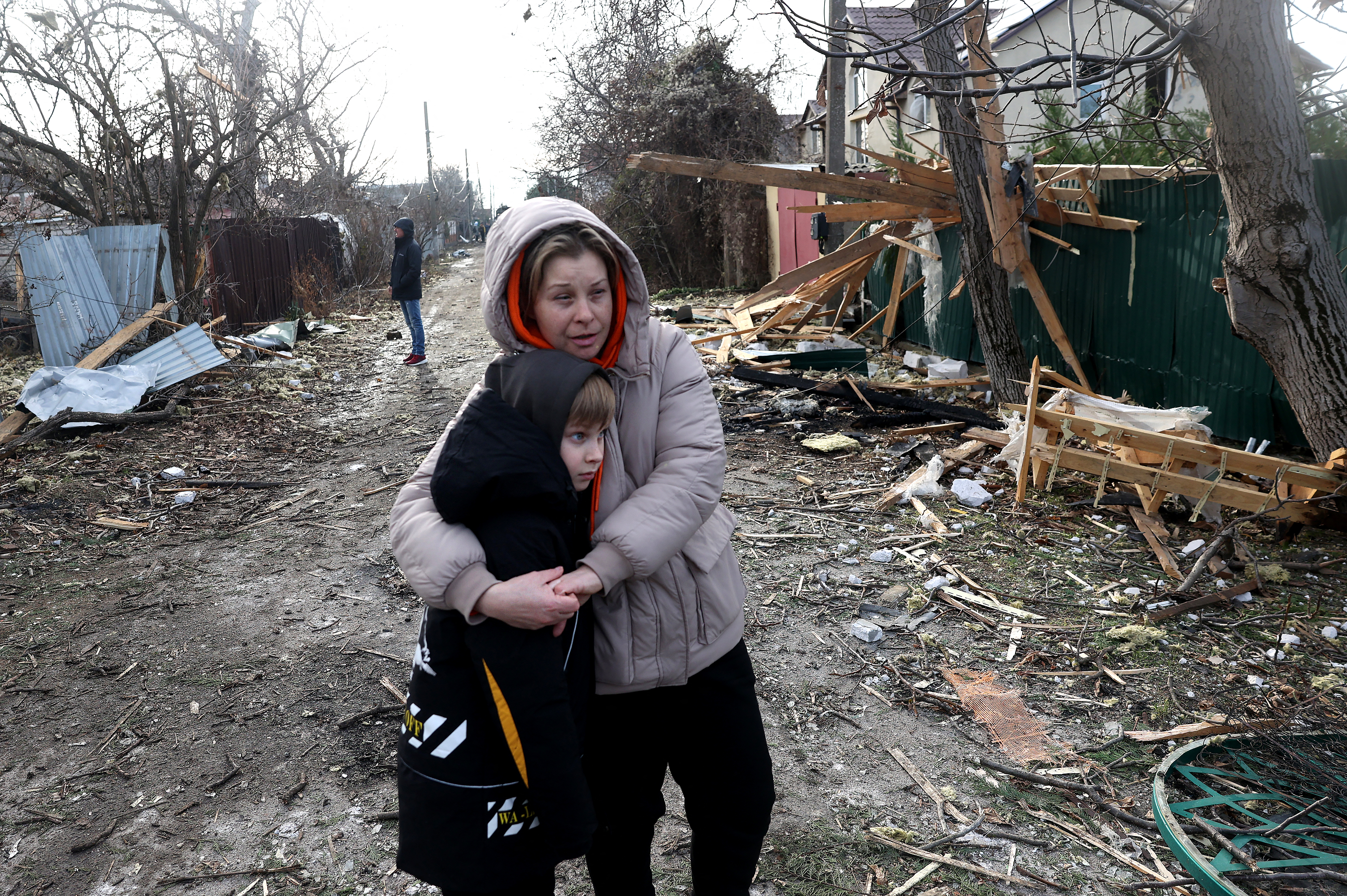 A woman holds a boy as they look at bombed out houses following a Russian attack. There's debris on the street behind them