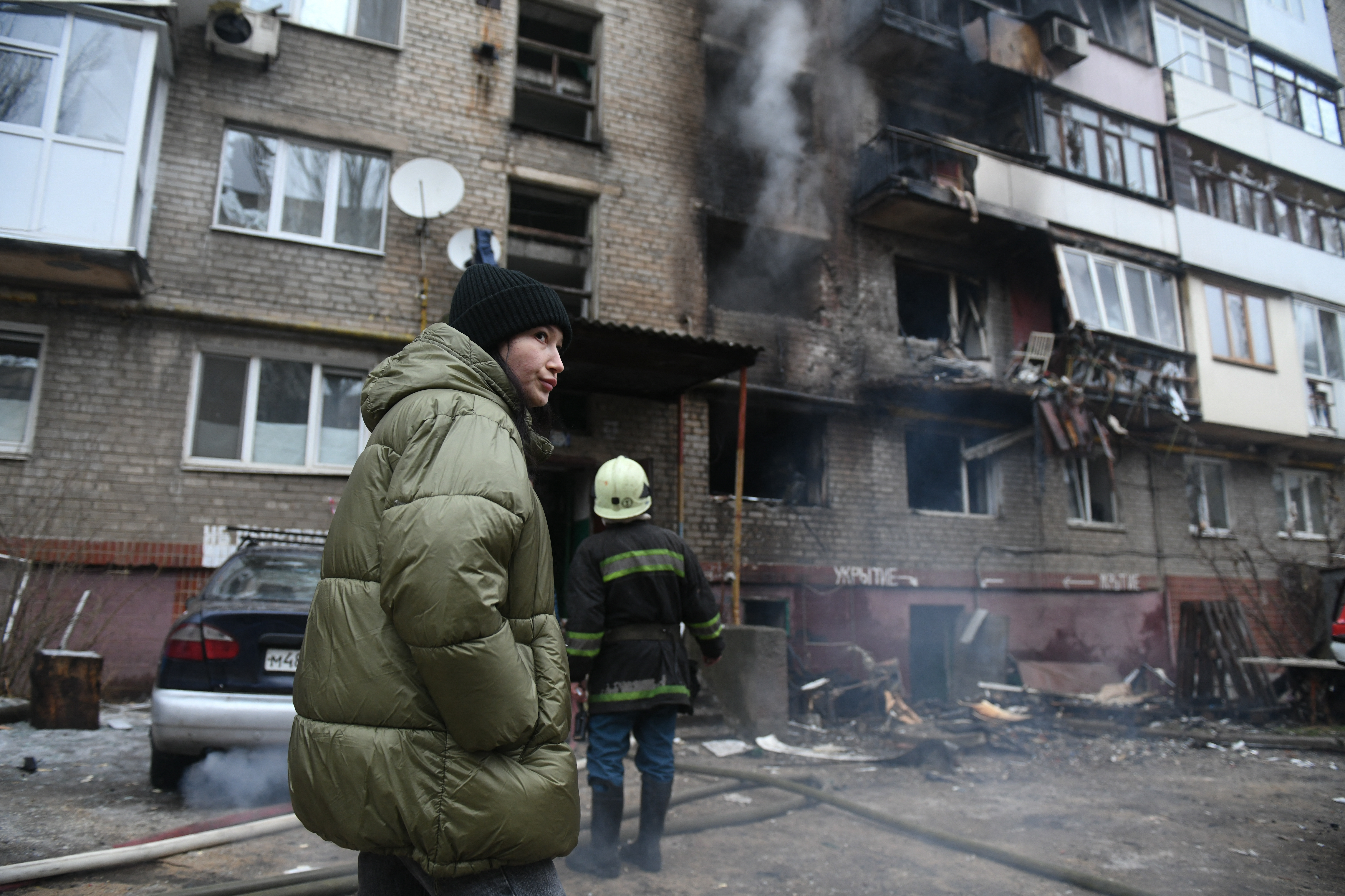A woman outside an apartment block hit by shelling in a region of eastern Donetsk occupied by Russia. She is wearing a puffa jacket and hat. Firefighters are working behind her.