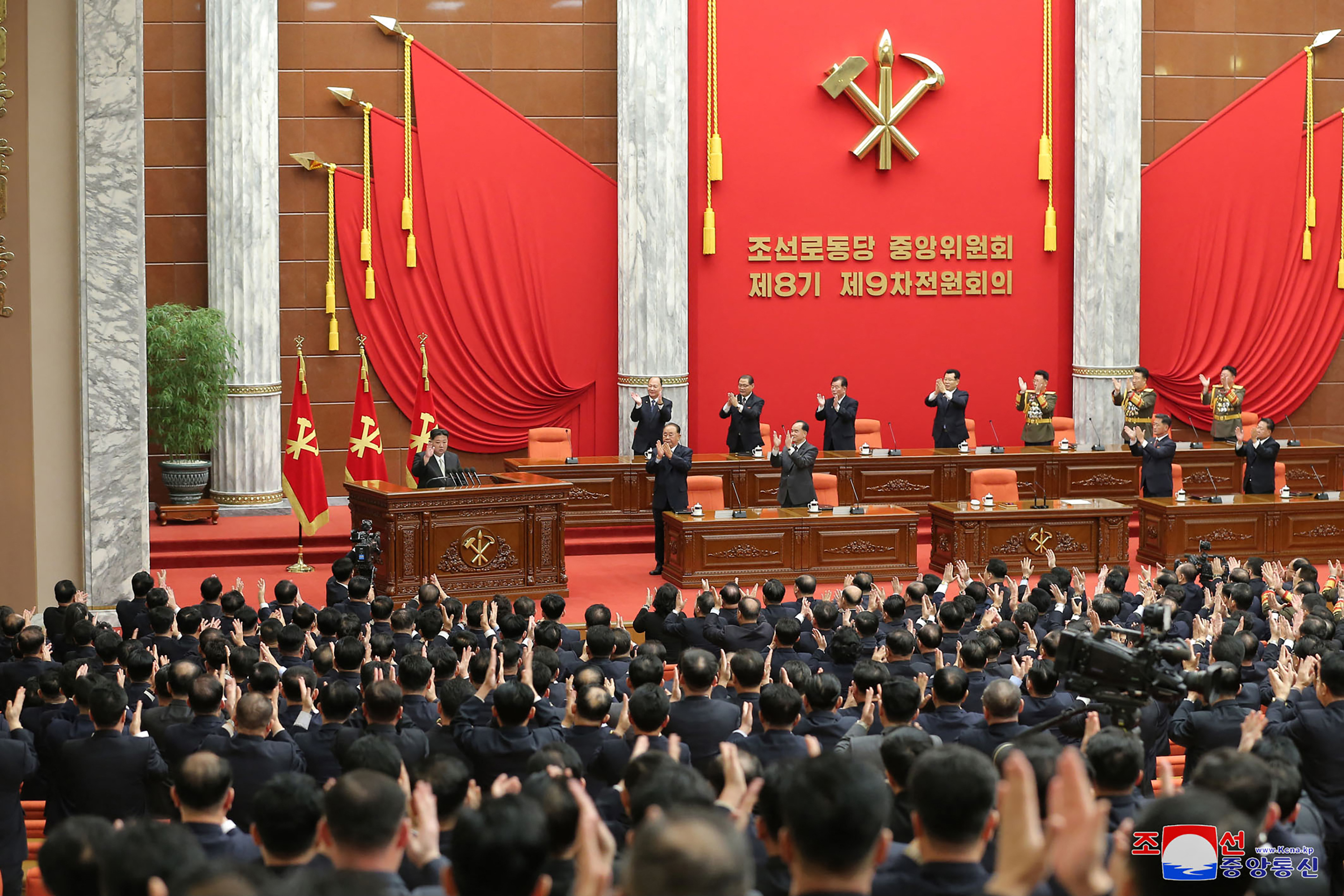 A view of the Workers' Party meeting. Kim Jong Un is on the left of the podium, he is being applauded by the others on the stage as well as the delegates in the hall The wall behind is decorated with red banners, party flags and the hammer and sickle.