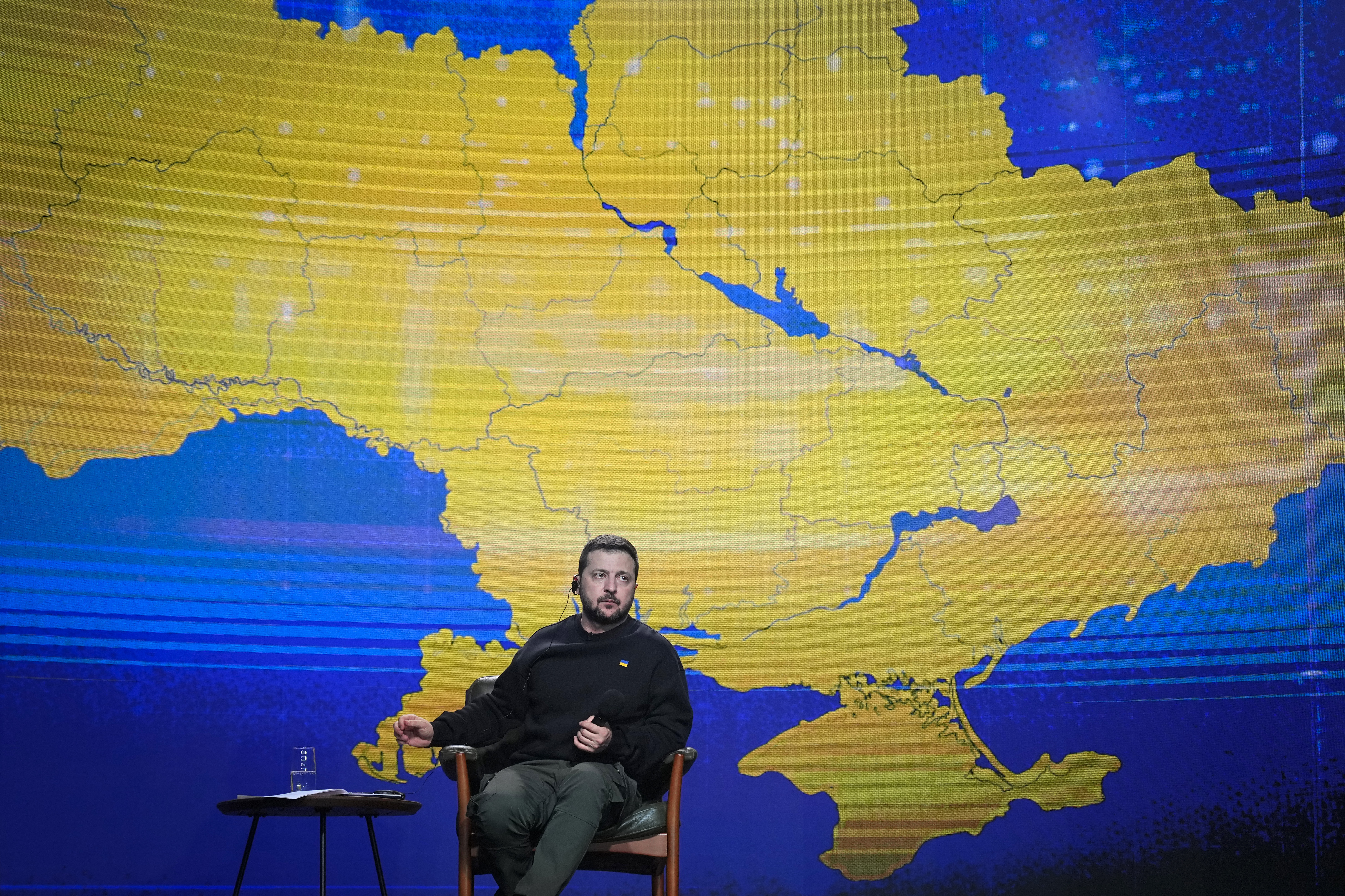 Volodymyr Zelenskyy sitting on stage during his annual press conference. A map of Ukraine is in blue and yellow behind him.