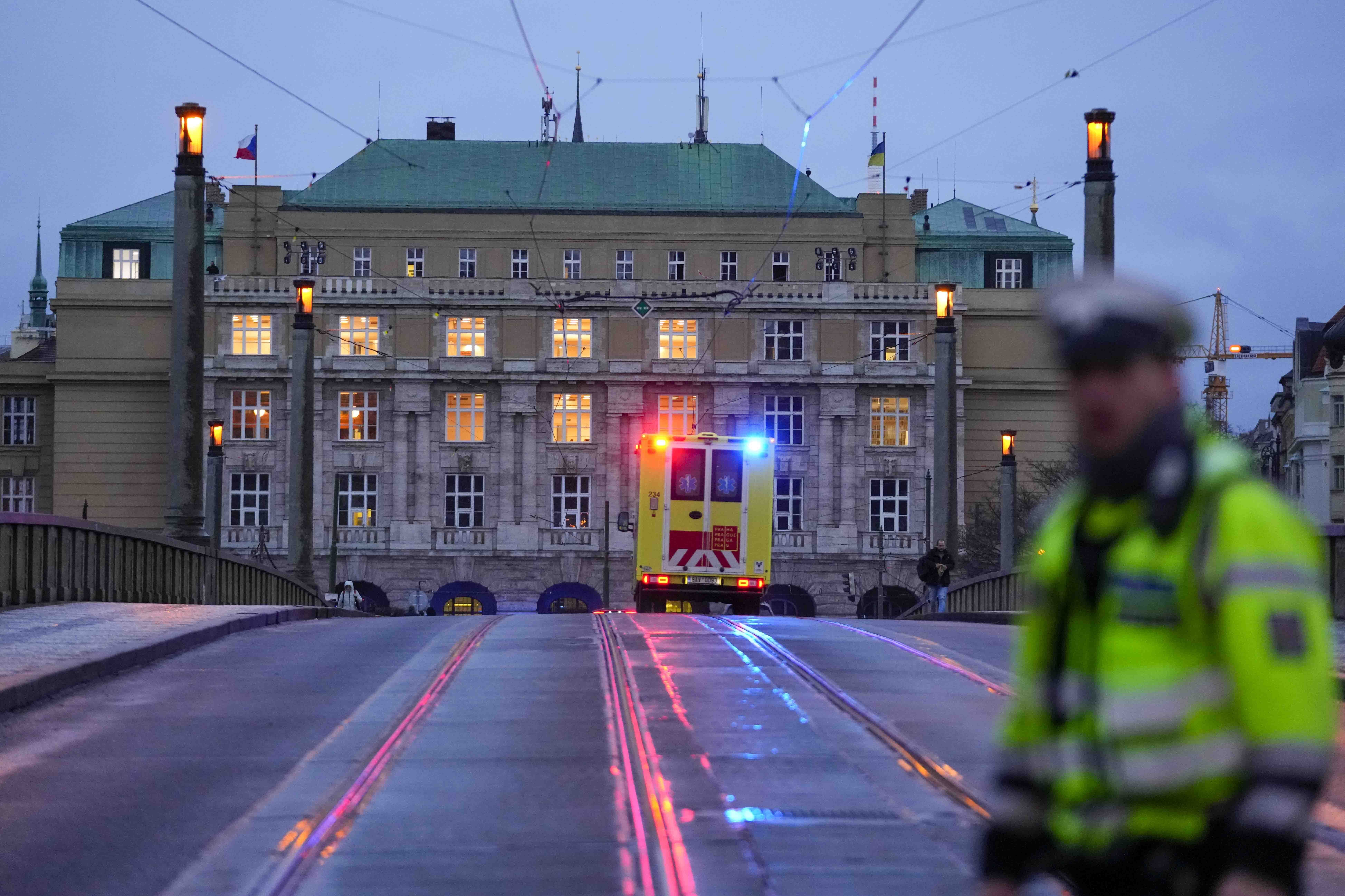 An ambulance with its lights flashing heading towards the Charles University. A police officer is in front. The building is behind.
