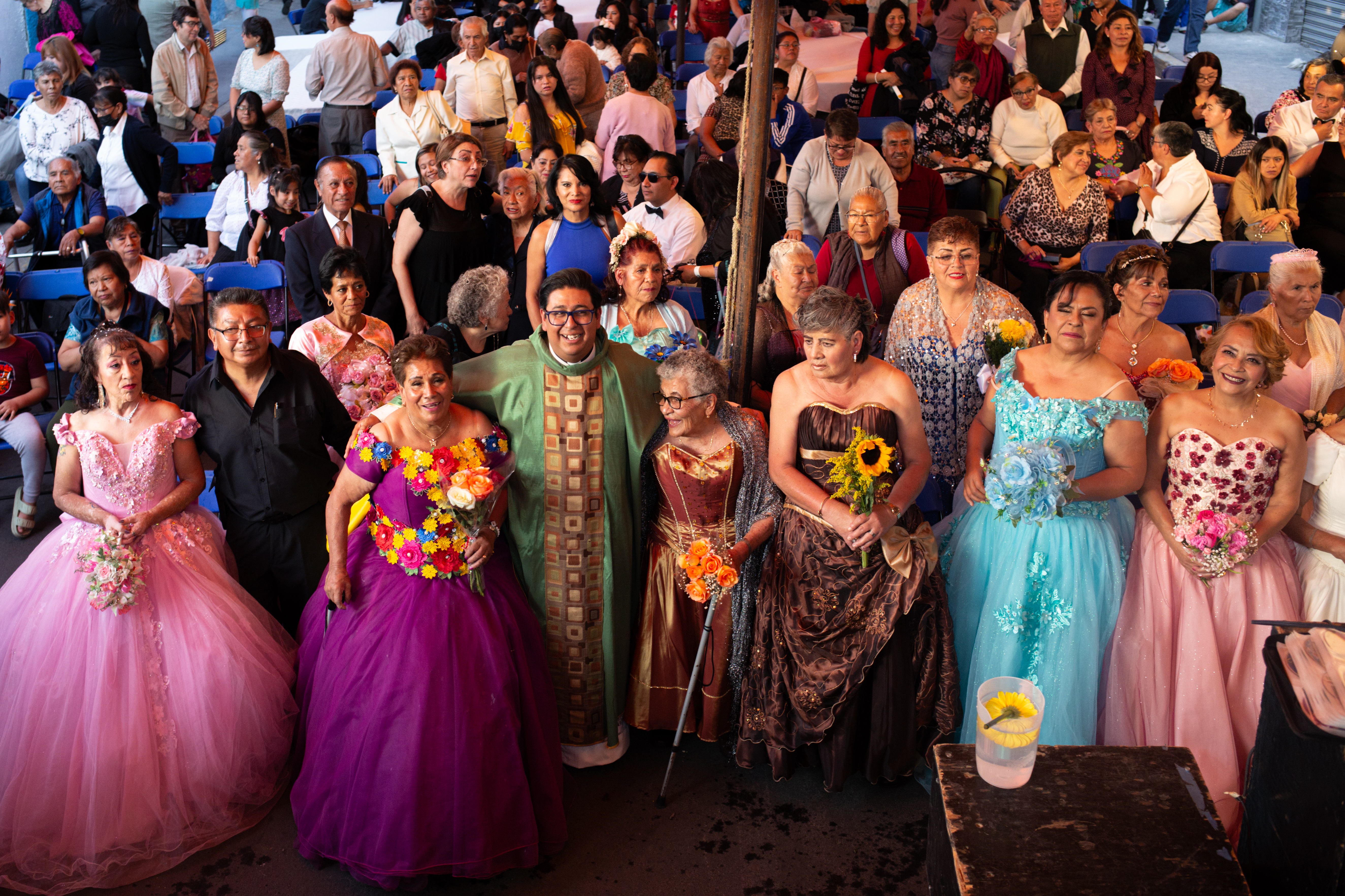 A priest in a green frock with gold trim stands amid a sea of women dressed in beautiful, multi-coloured ballgowns.